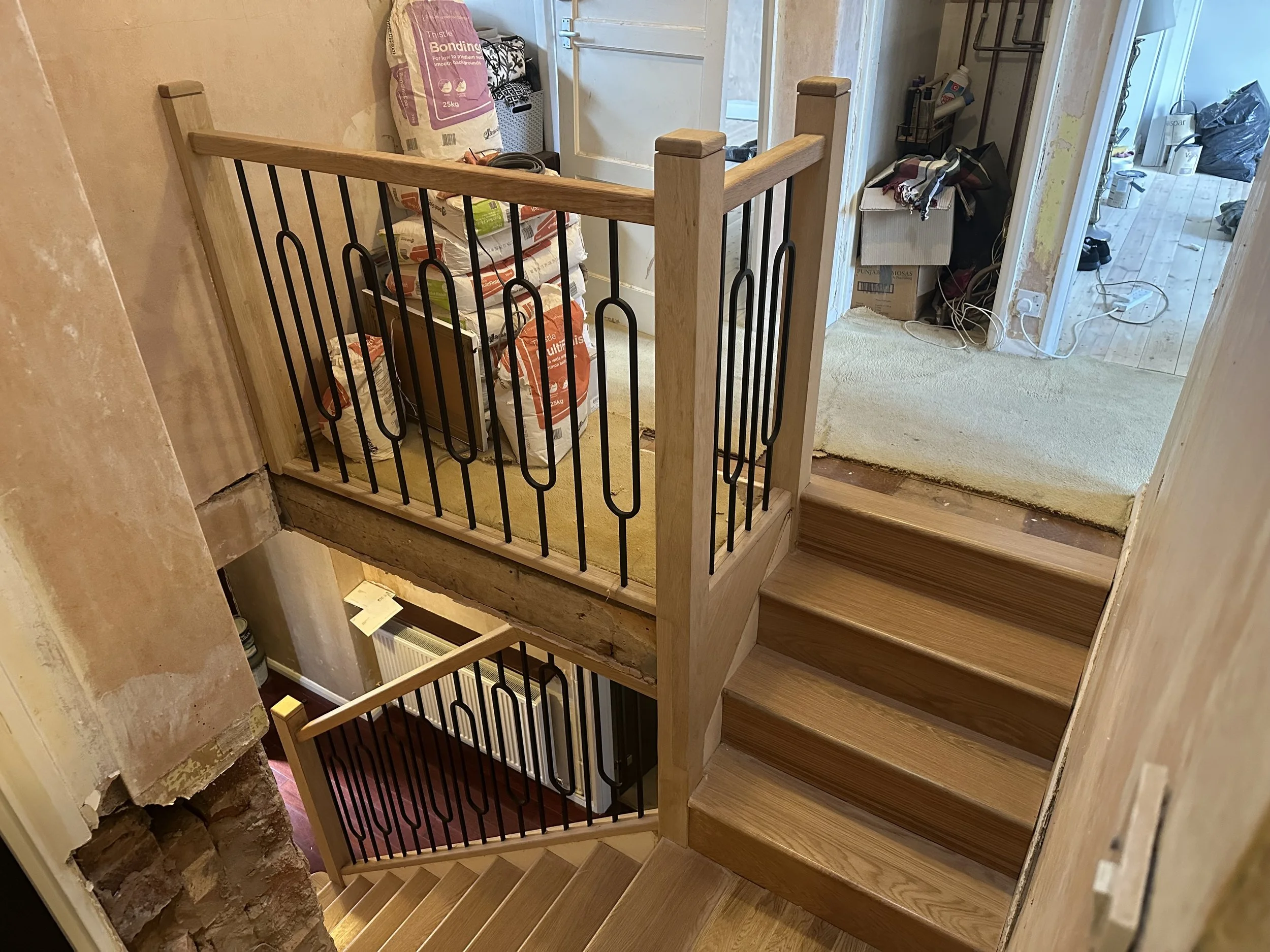 View of a staircase with wooden steps and black metal railings, leading to a second floor. The staircase is in the middle of a room, with a door at the top, and a wall with exposed bricks on the left. The nearby room has visible clutter including bag