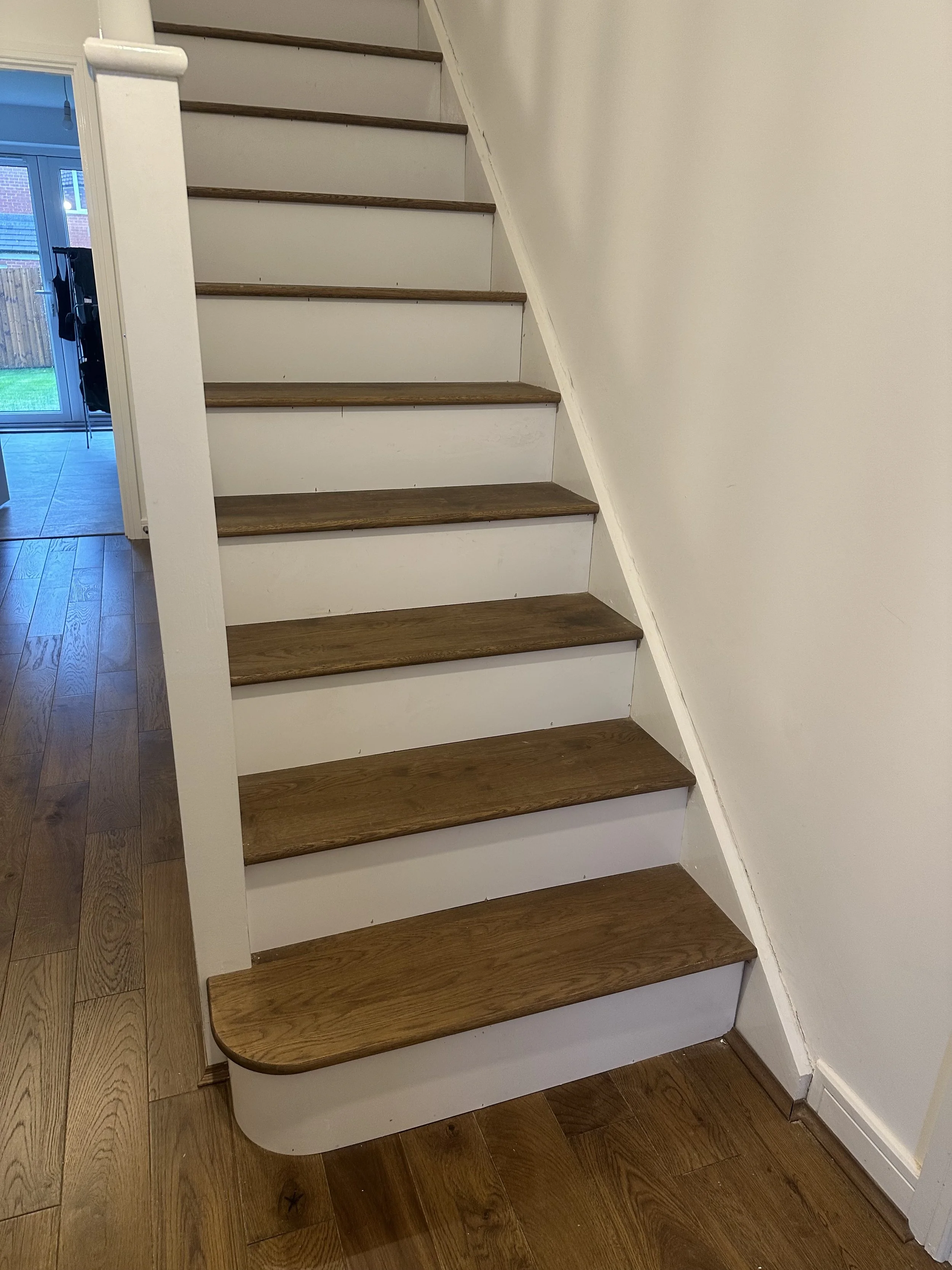 Wooden staircase with white sides and a rounded bottom step, inside a home with hardwood floors and white walls.