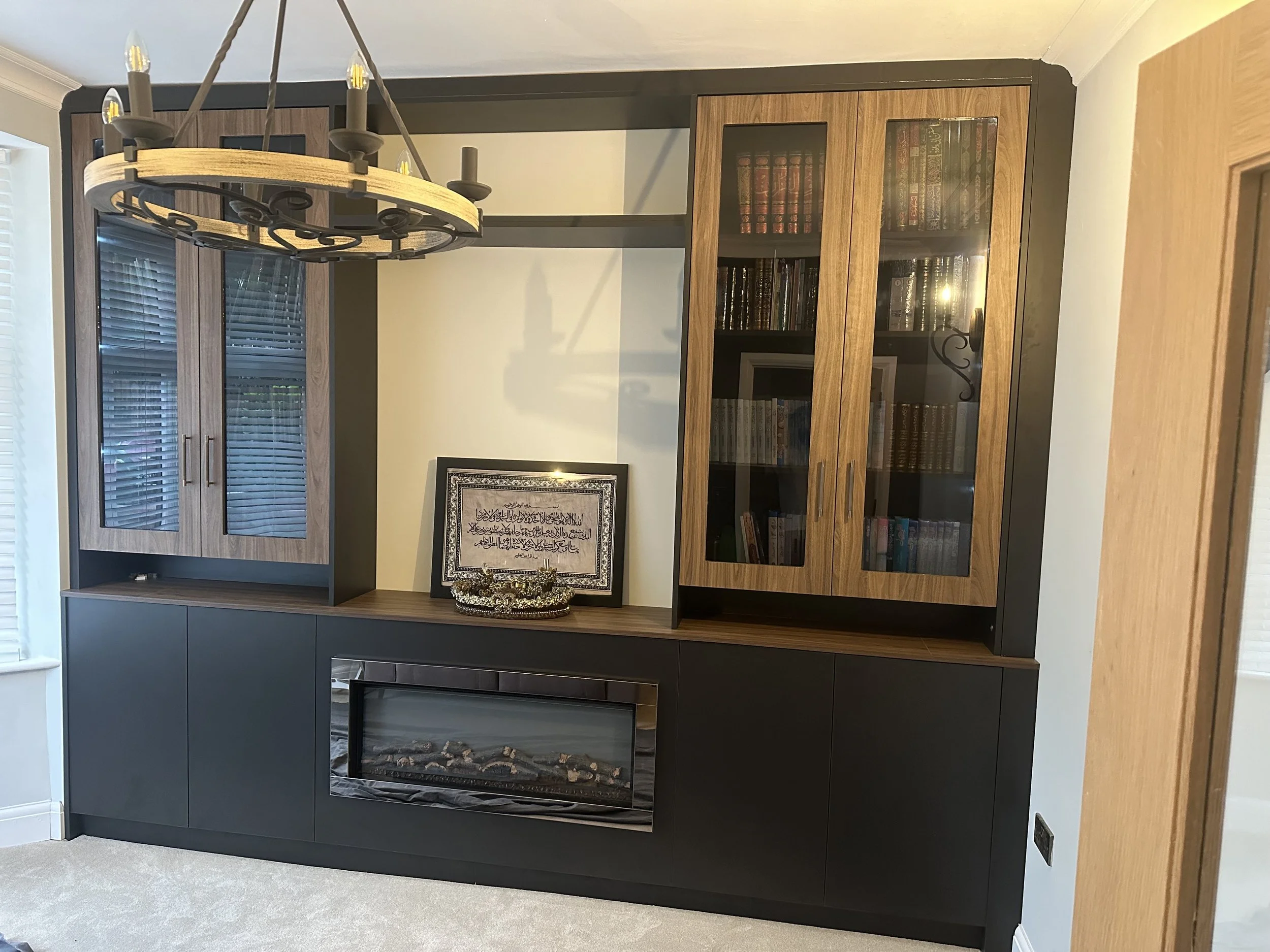 A modern black and wood-colored entertainment unit with a built-in electric fireplace at the bottom, glass-fronted cabinets filled with books, a framed Arabic calligraphy art piece, a decorative tray, and a chandelier hanging above.