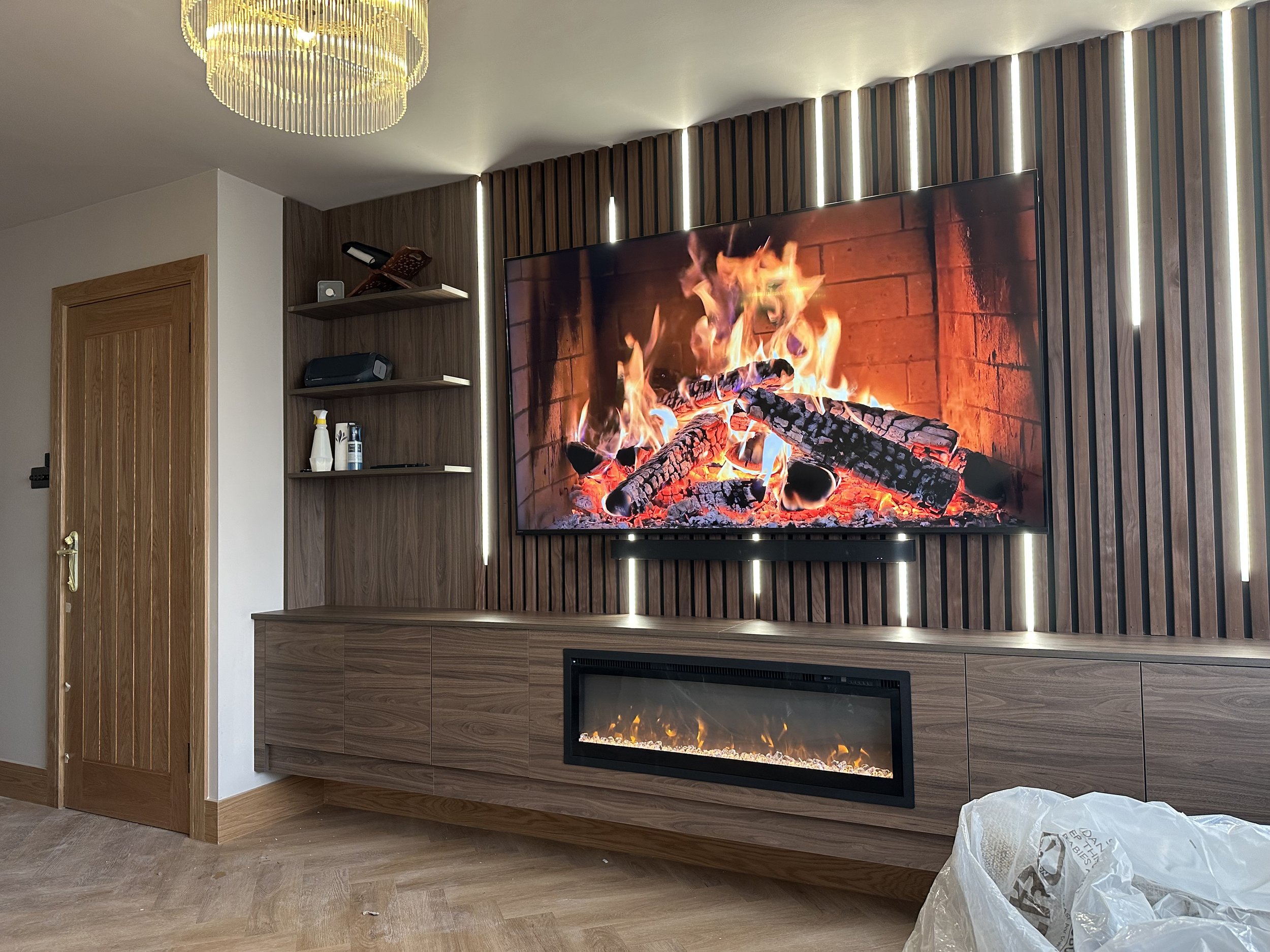 Living room with a wall-mounted flat-screen TV displaying a realistic fireplace scene and a modern electric fireplace below it. The wall features vertical wooden paneling with LED accents. A chandelier hangs from the ceiling, and there are shelves with electronic devices and cleaning supplies on the left side of the image.