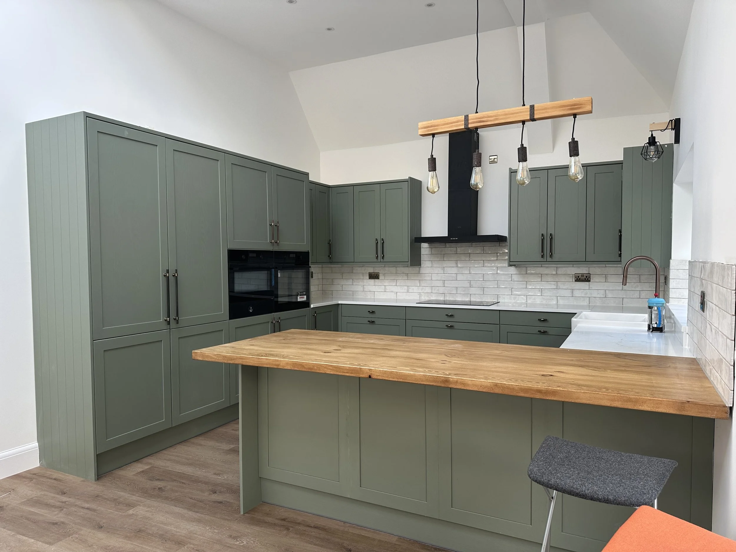 Modern kitchen with green cabinets, wooden countertop, white brick backsplash, hanging light fixtures with Edison bulbs, and a small kitchen stool.