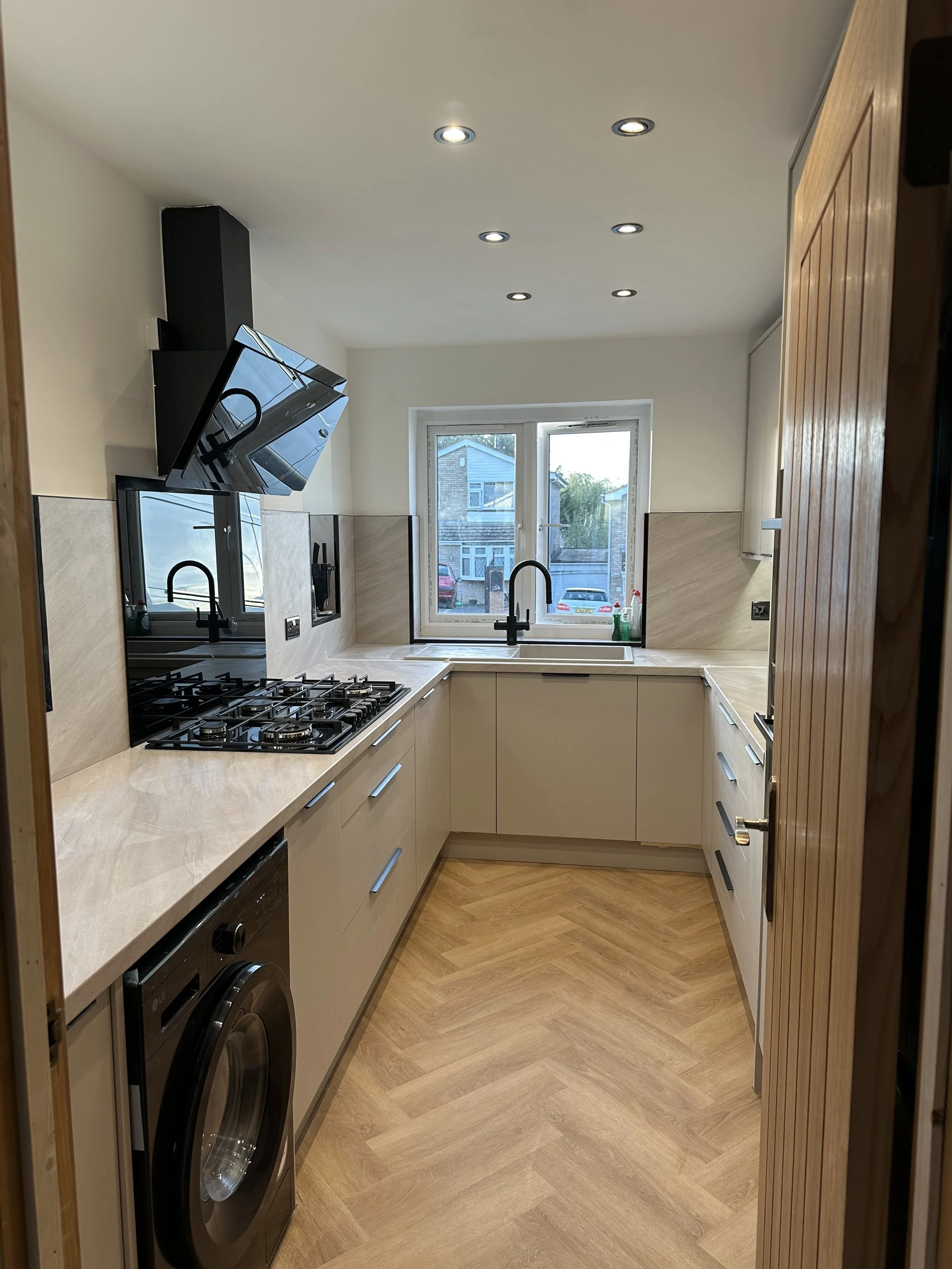 A modern kitchen with beige cabinets, a beige countertop, a window above the sink, four recessed ceiling lights, a gas stove, a washing machine, and a wooden door.
