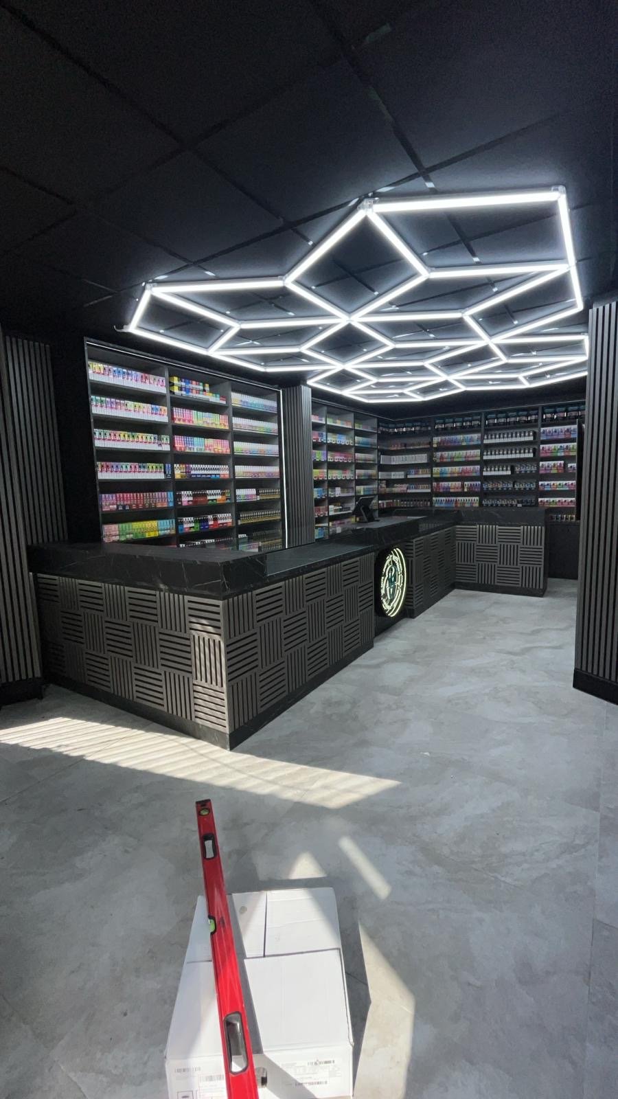 Interior of a modern vape shop with black paneling, illuminated geometric ceiling lights, and shelves filled with colorful vape bottles. A counter with a black marble top and a logo on the front. Tools and supplies are set on the counter, and a level tool is placed on a white box on the floor.