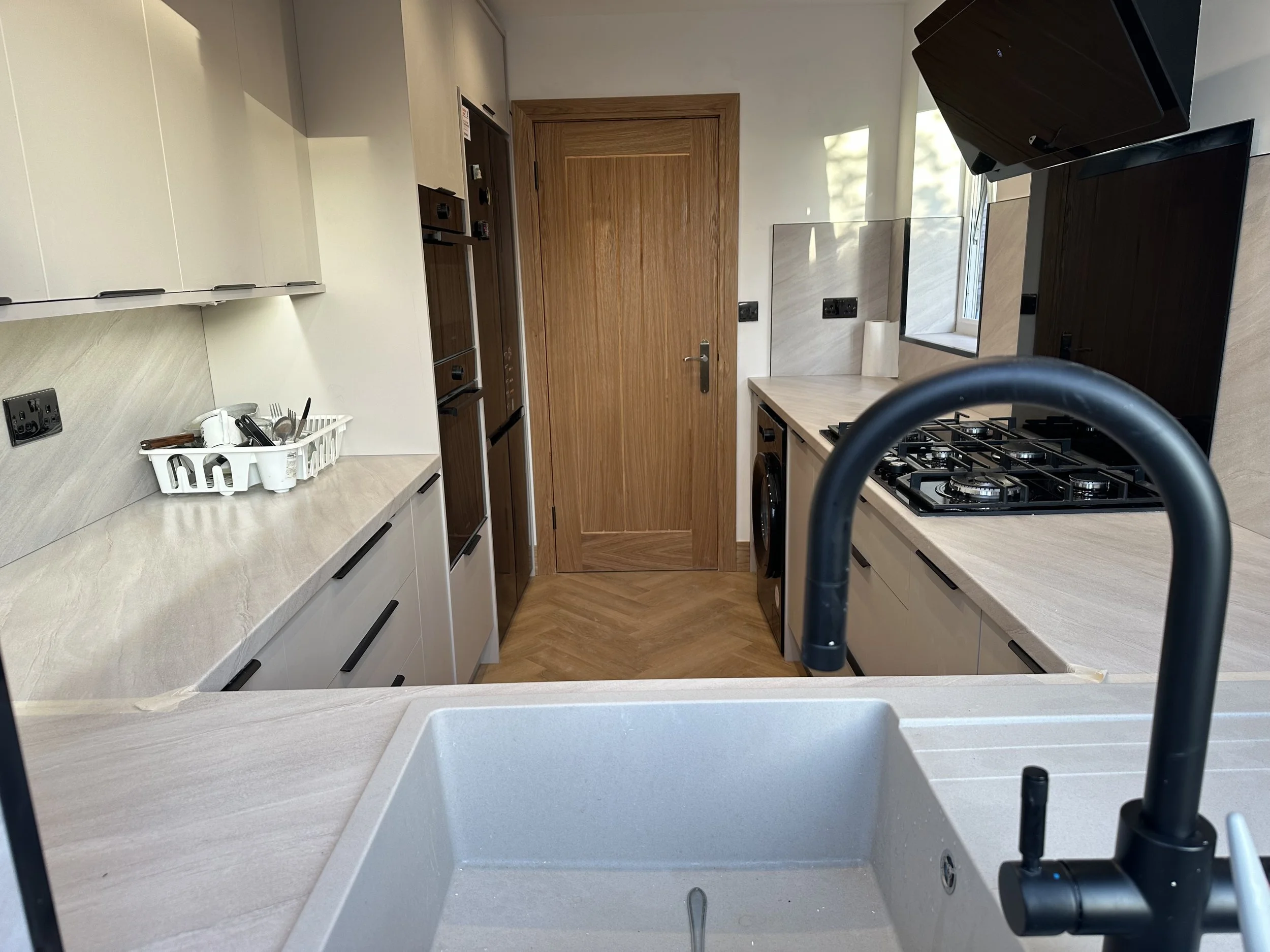A modern kitchen with white cabinets, a wooden door, a gas stove, a window, and a white sink with a black faucet in the foreground.