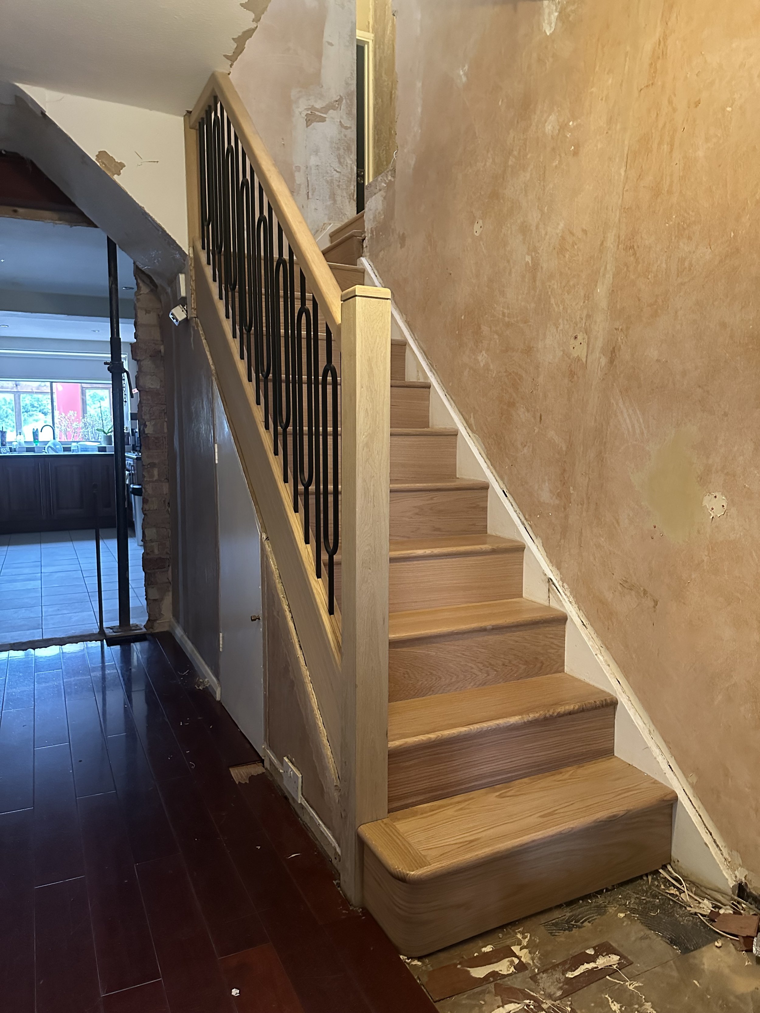 Interior stairway undergoing renovation with missing wall sections, a new wooden staircase with black metal railing, and construction debris on the floor.