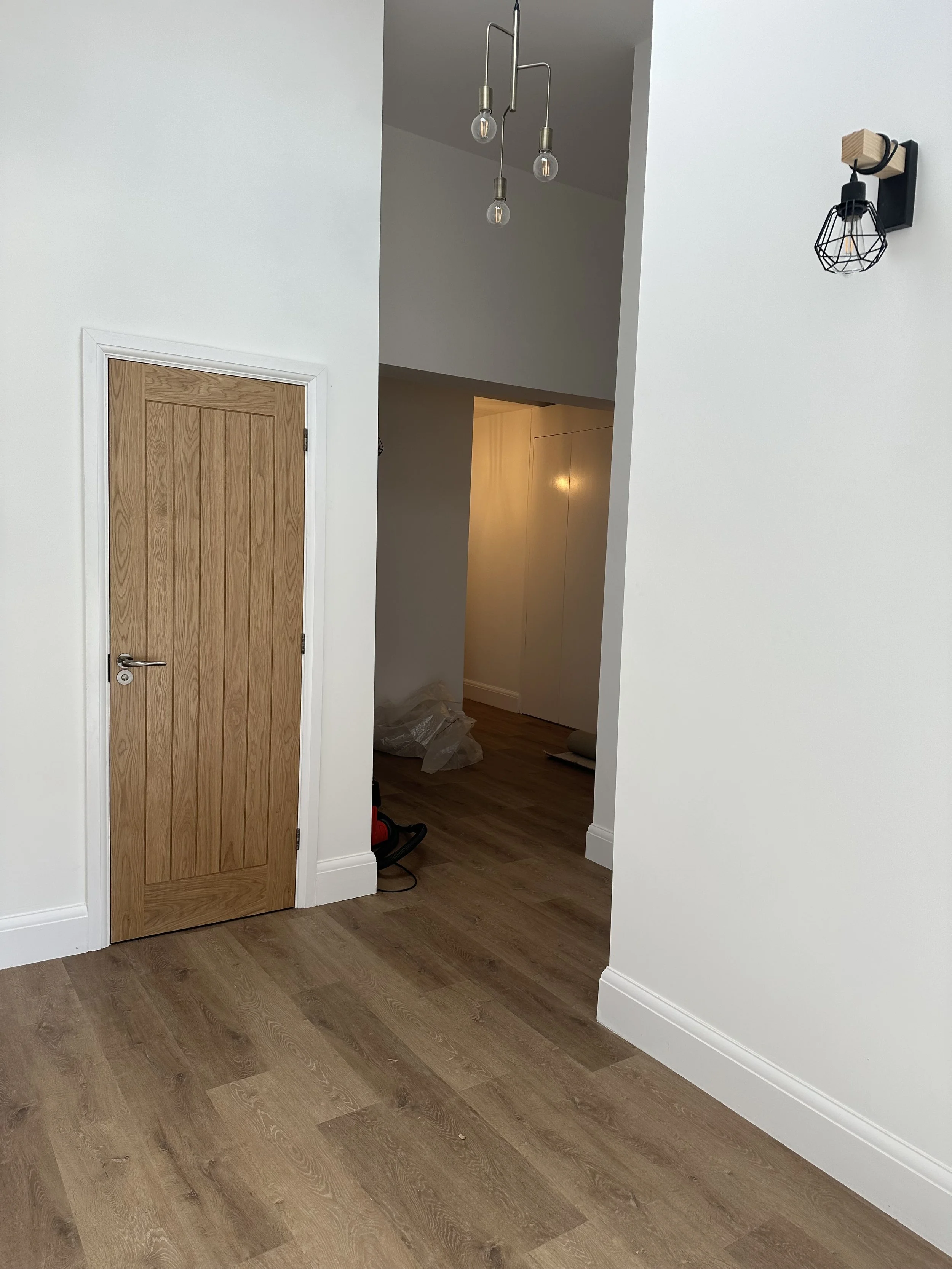 Empty room with wooden floors, white walls, a wooden door, a hanging light fixture with three bulbs, and a black wall light fixture. There are some plastic bags and a vacuum cleaner on the floor in the background.