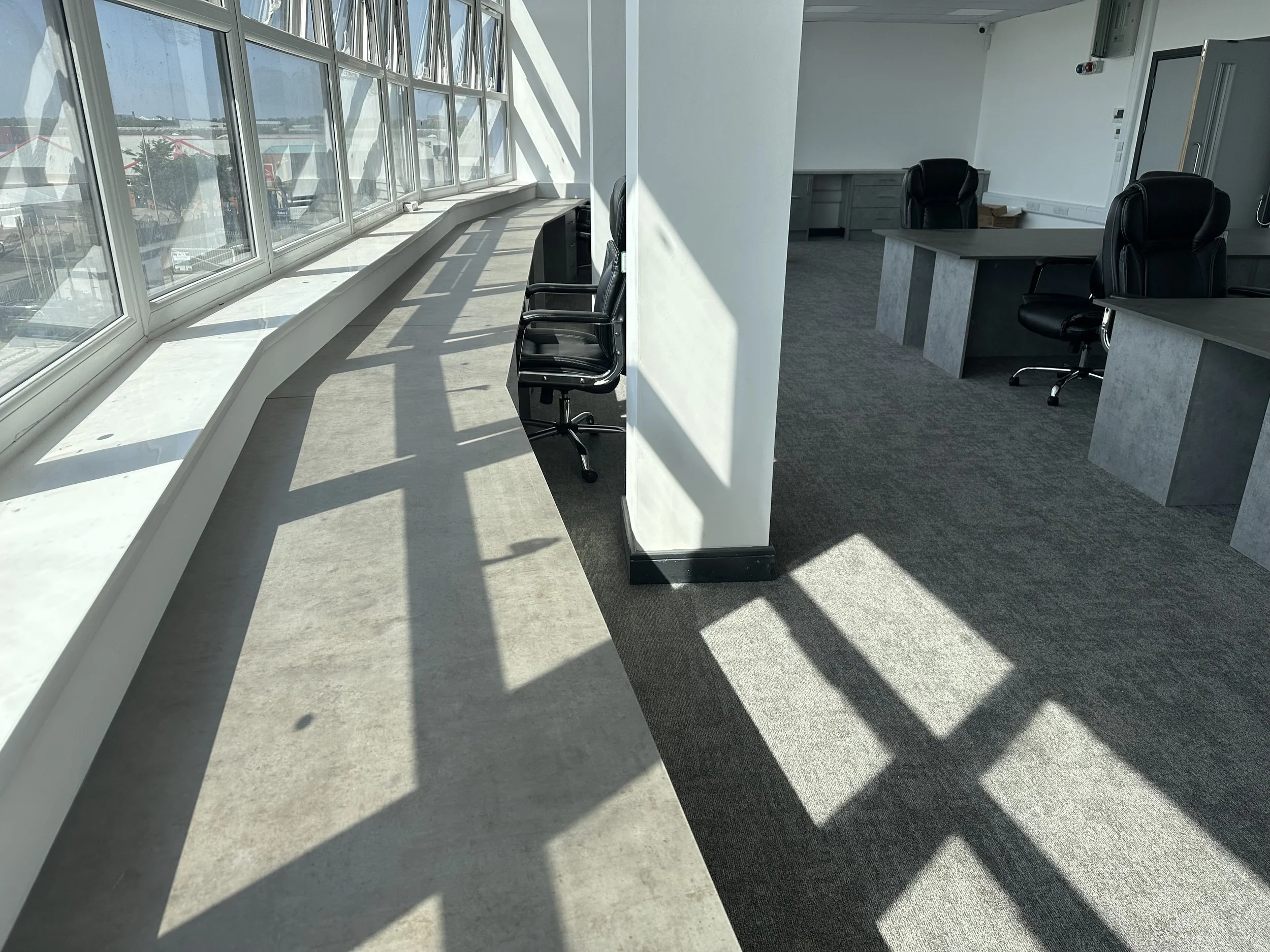 An office space with large windows on the left, black office chairs, desks, and a carpeted floor.