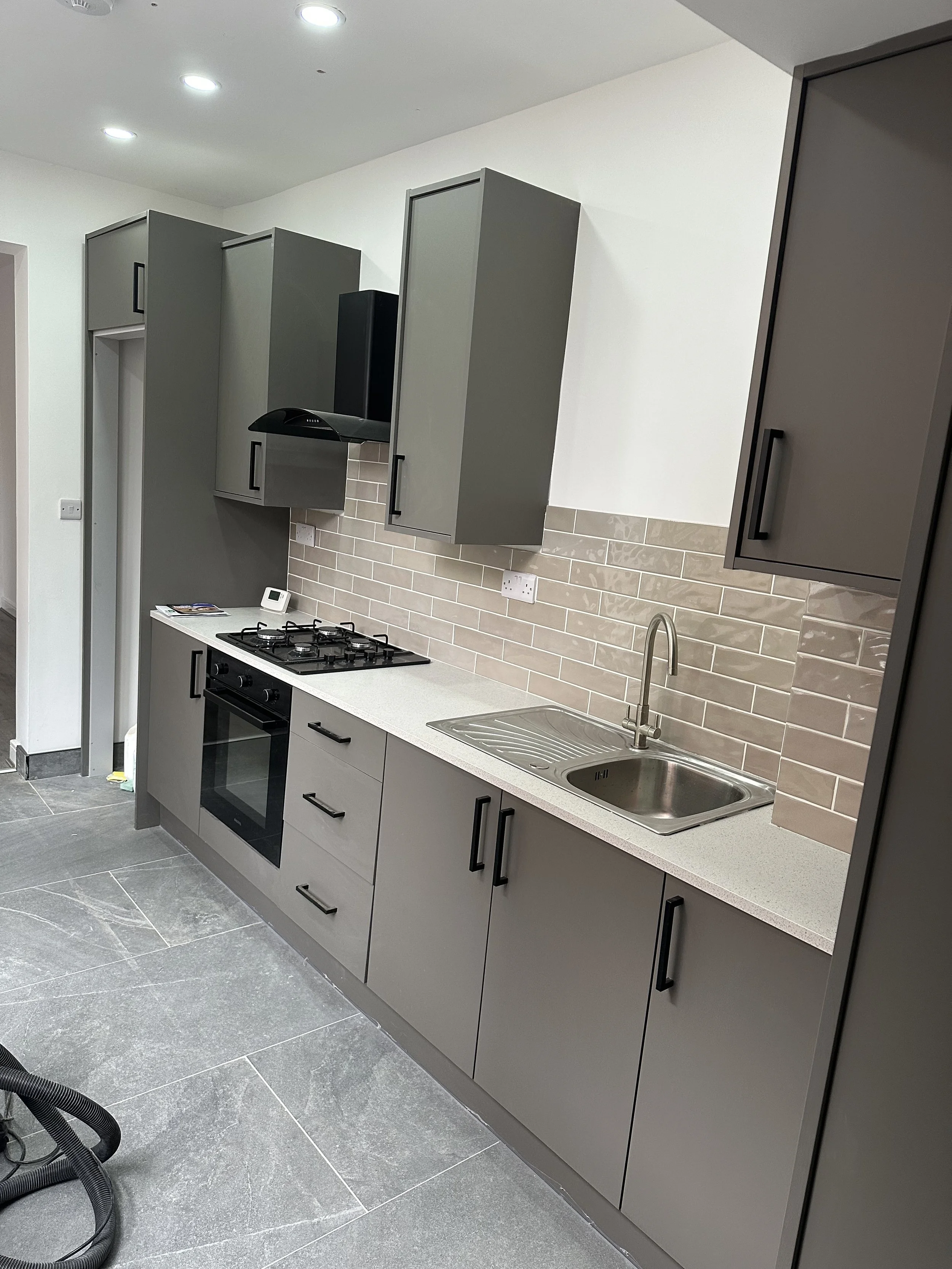 Modern kitchen with grey cabinetry, beige tiled backsplash, stainless steel sink, and grey floor tiles. There is a built-in oven and gas cooktop, and a small digital timer on the countertop.