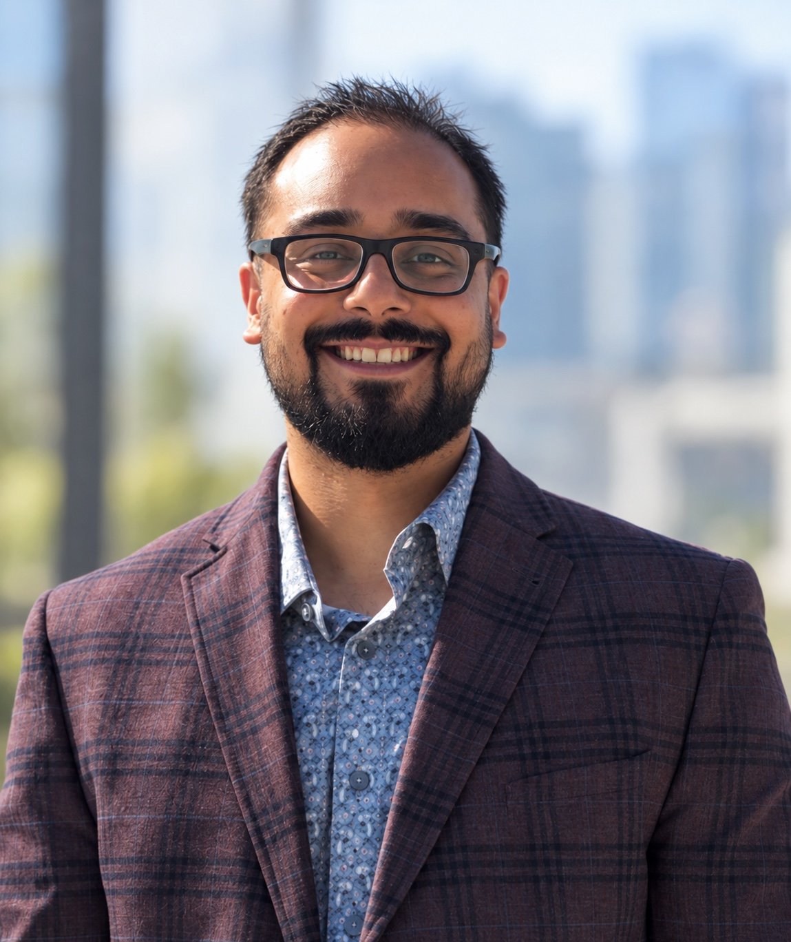 Smiling man wearing glasses and a suit, outdoors with a blurred cityscape background.