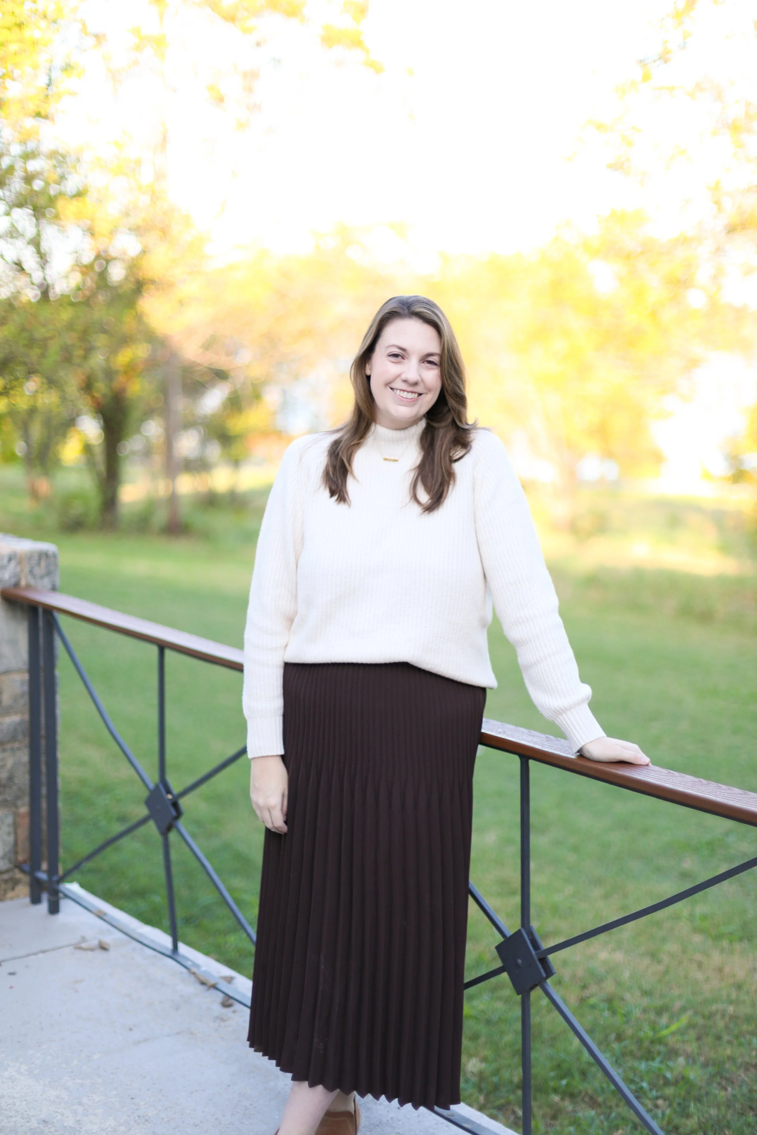 A young woman with long brown hair smiling outdoors, wearing a cream sweater and a dark pleated skirt, standing near a black metal fence on a concrete path, with a background of yellow autumn leaves and trees.