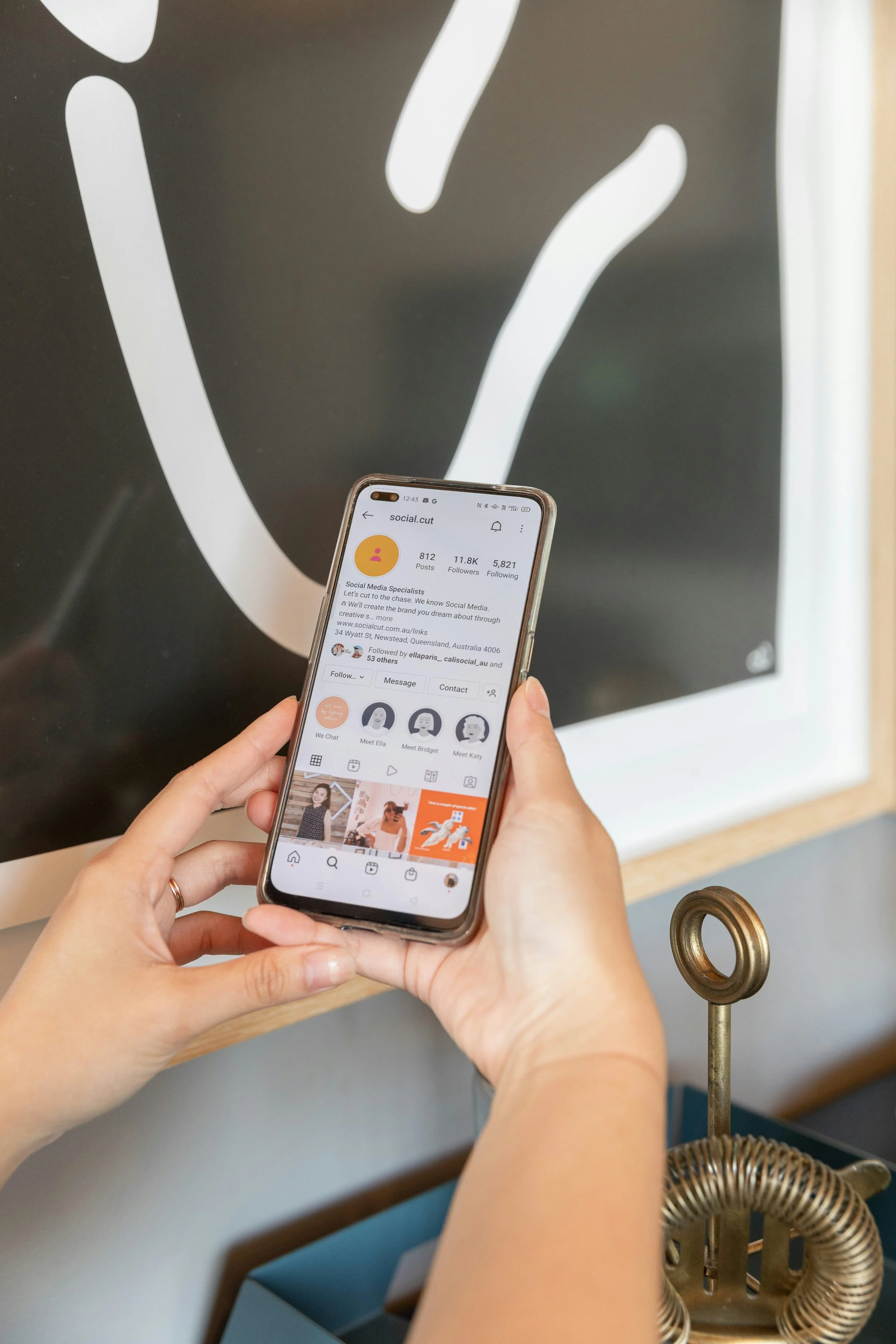 Person holding a smartphone displaying an Instagram profile for 'social.cut' with 812 posts, 11.8K followers, and 5,821 following, in front of a black and white design on a wall.