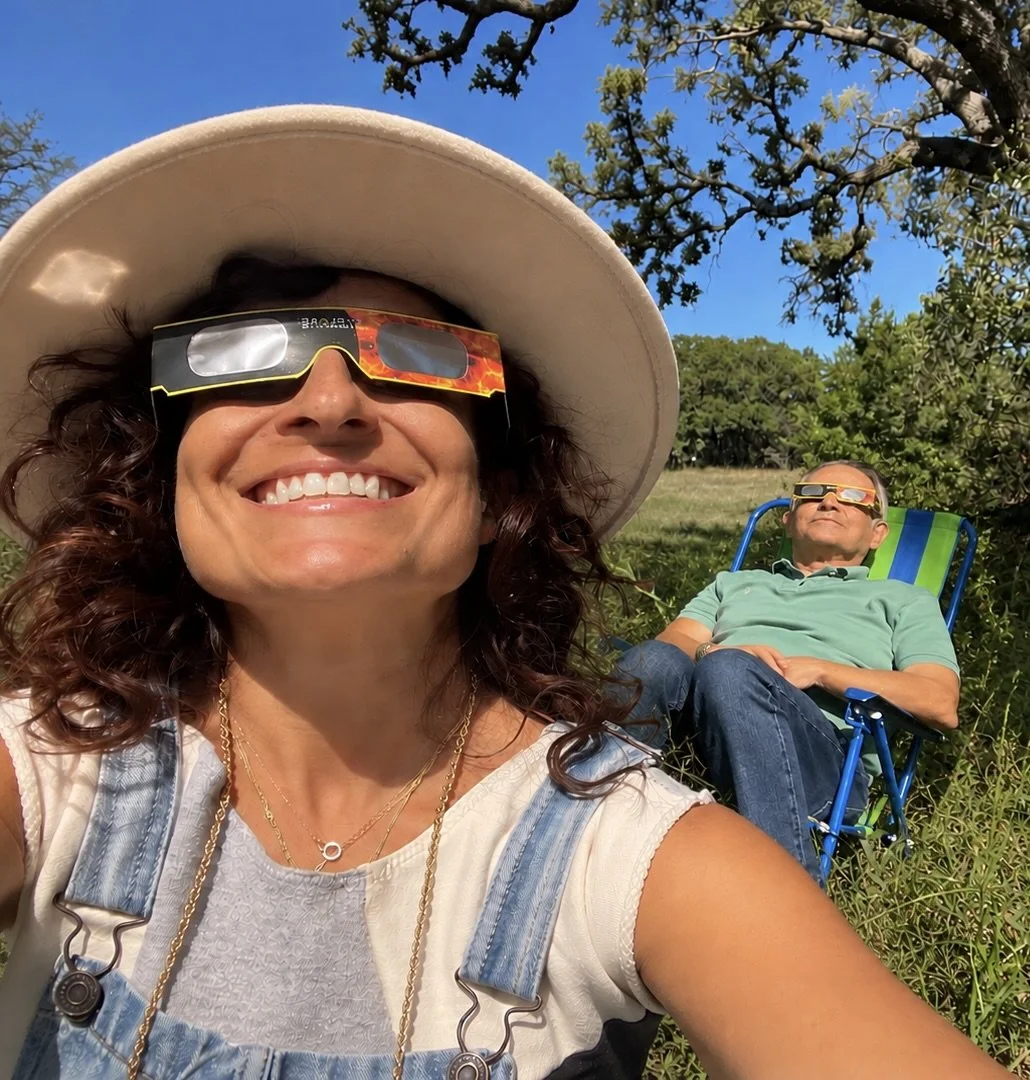 Smiling woman wearing eclipse glasses and a sun hat takes a selfie outdoors while a man relaxes in a lawn chair behind her.