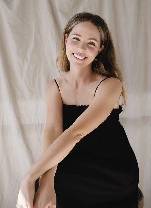 Smiling pregnant woman in a black spaghetti-strap dress posing against a cream fabric backdrop