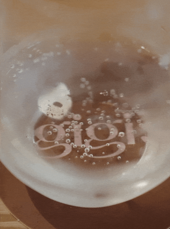 Close-up of a sparkling water with bubbles in a glass on a wooden table with the logo for Gigi's Restaurant peeking out