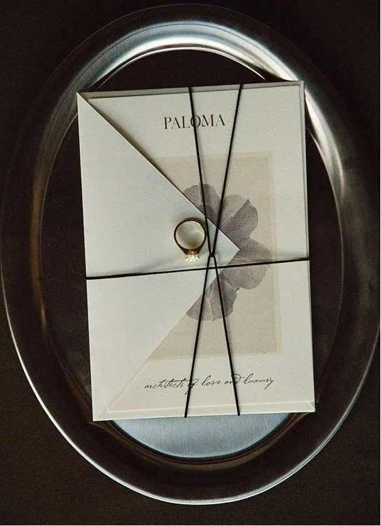 Elegant Paloma invitation tied with black cord and gold ring, displayed on an oval reflective tray