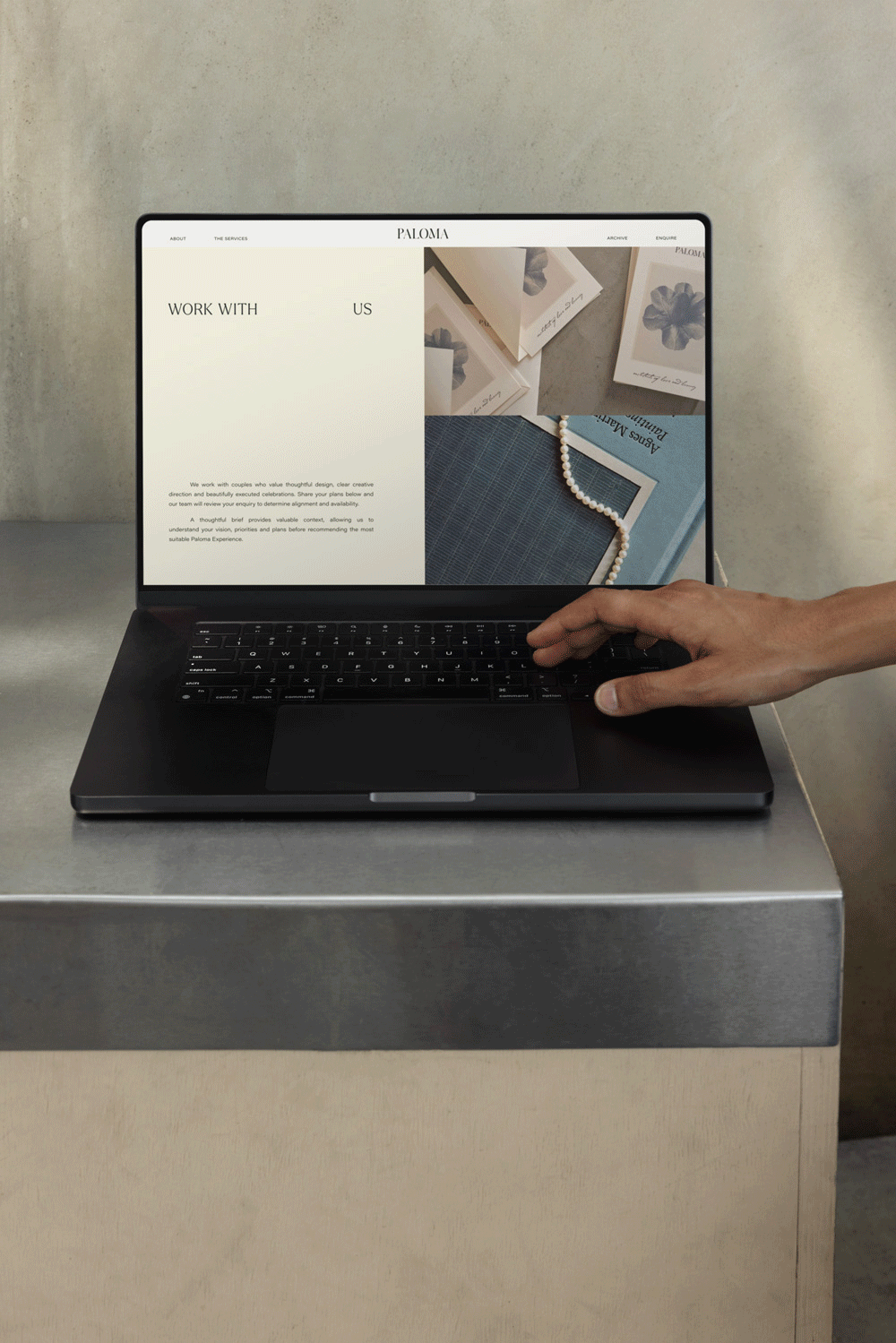 Hand using a black laptop displaying a Paloma creative design studio website on a gray metal table