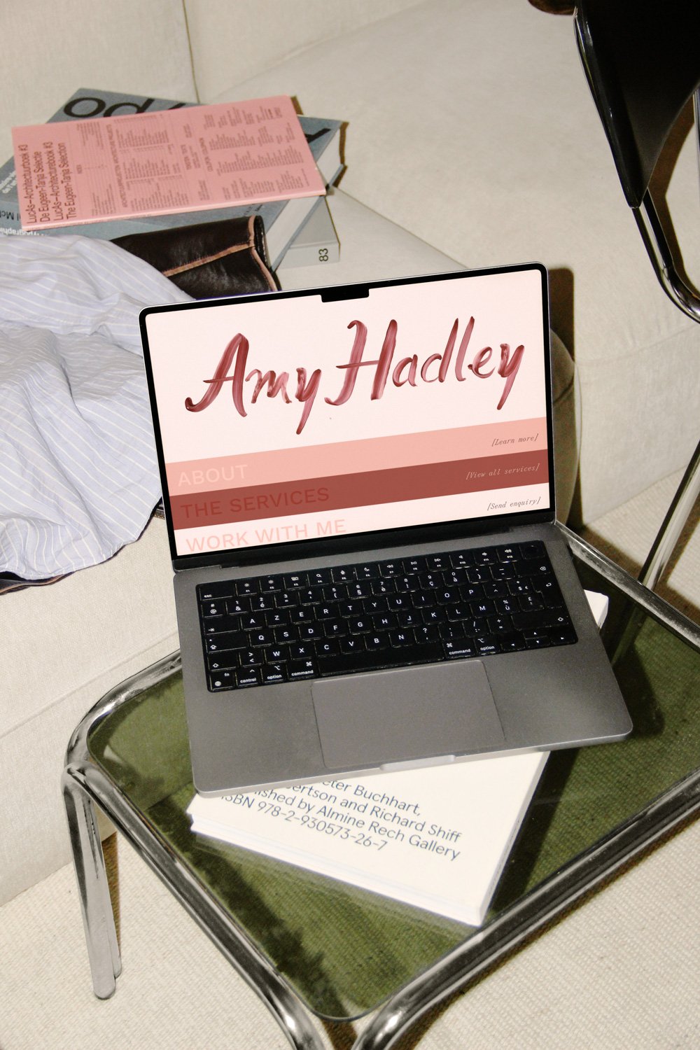 MacBook displaying an Amy Hadley portfolio website on a chair beside books and a pink pamphlet