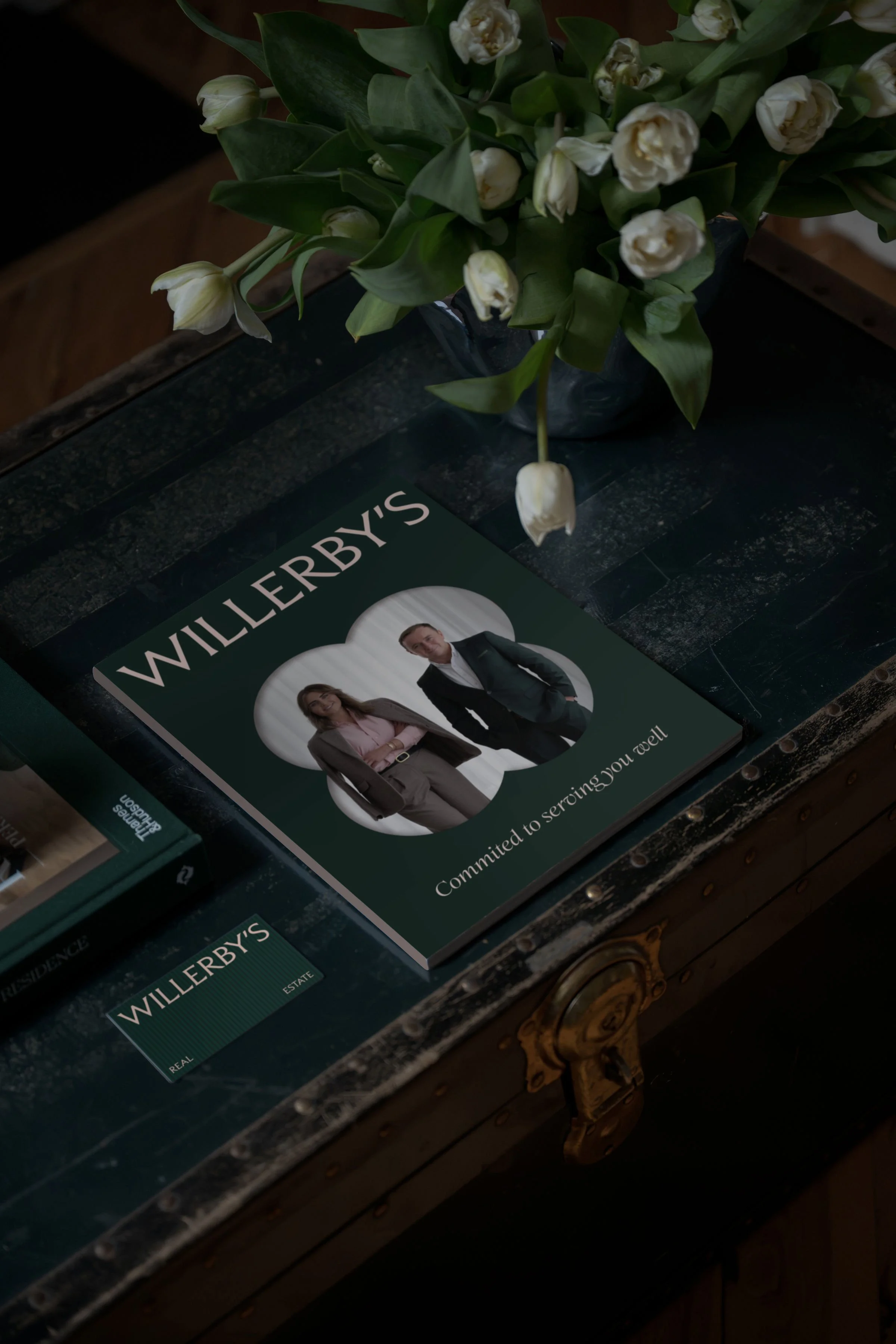 White flowers and green leaves arranged beside a dark green Will Ferrell book on an antique wooden table