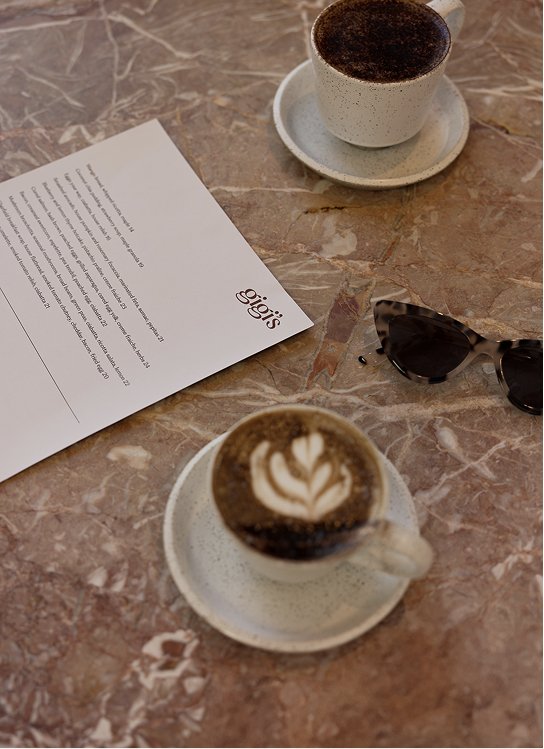 Two cappuccinos on saucers, a café menu, and sunglasses on a marble table
