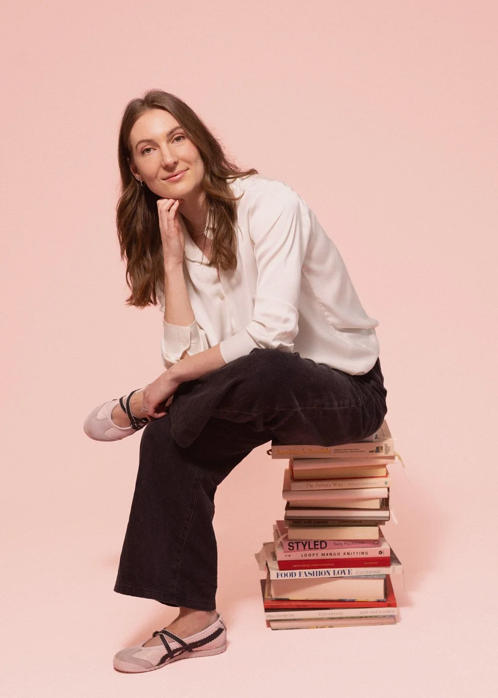 Woman in a white blouse and black pants sitting on a stack of books against a pink background