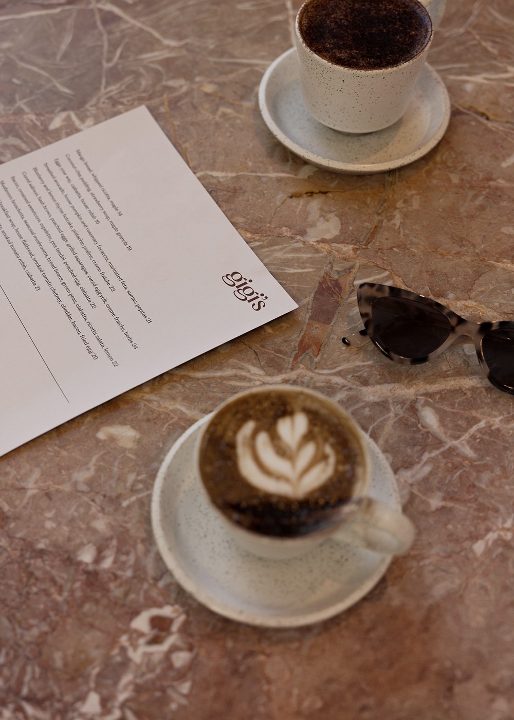 Coffee and cappuccino on saucers beside a cafe menu and sunglasses on a marble table