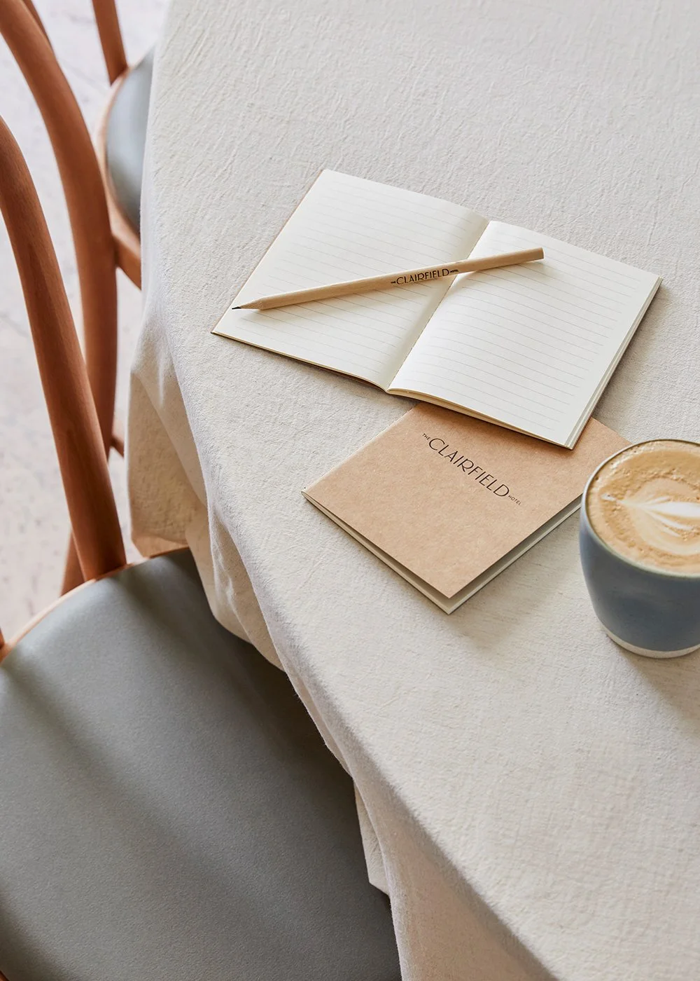 Open lined notebook with a pencil beside a Clairefield hotel booklet and latte on a white tablecloth