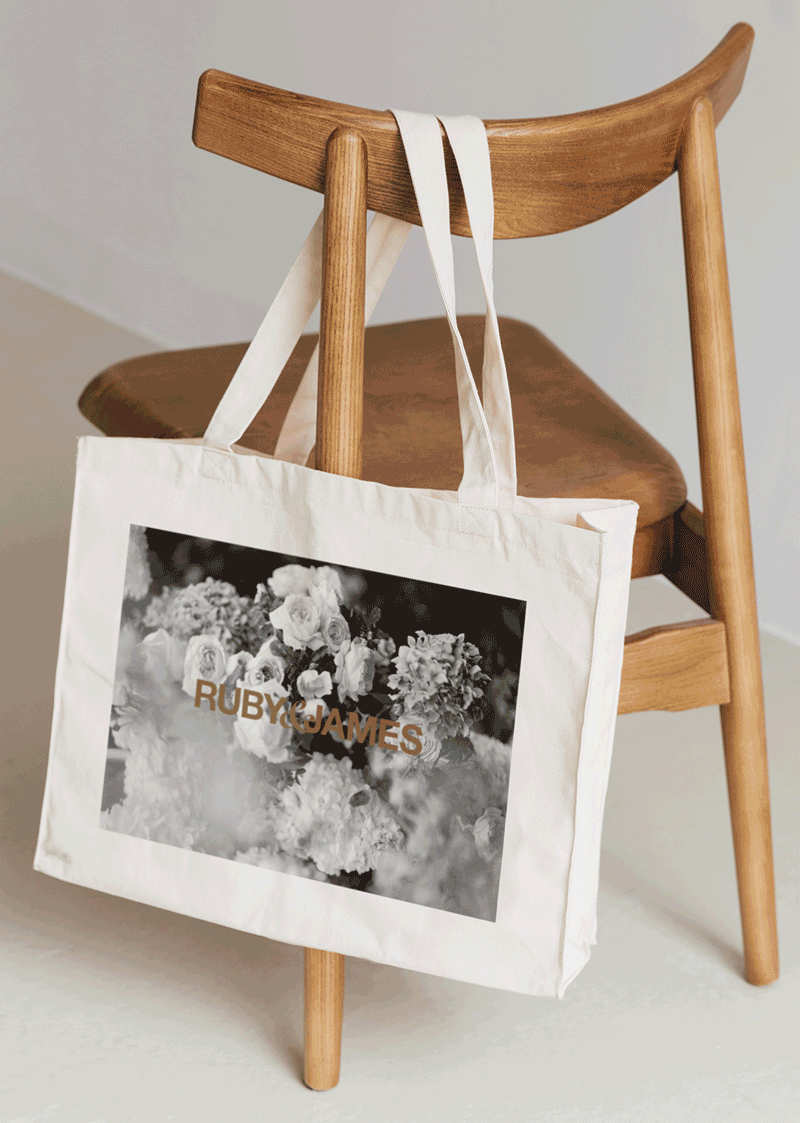 White canvas tote bag with a black-and-white floral print and “RUBY JAMES” text hanging from a wooden chair