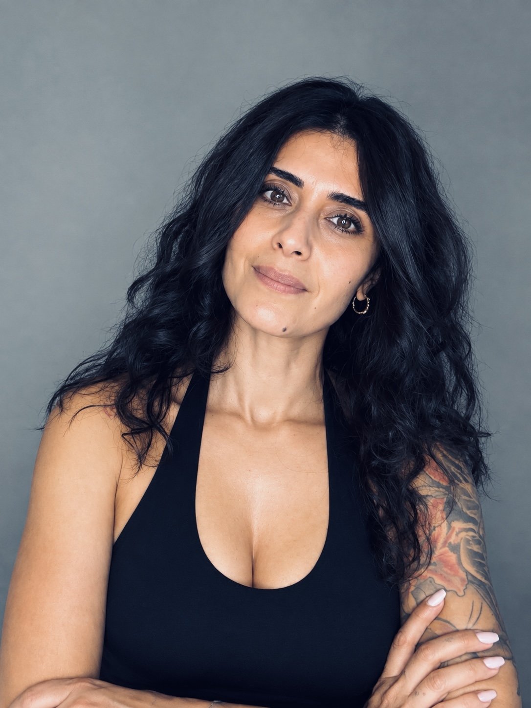Porträt einer Frau mit langen schwarzen Haaren, schwarzem Oberteil, Ohrring und farbigem Tattoo am Arm vor grauem Hintergrund