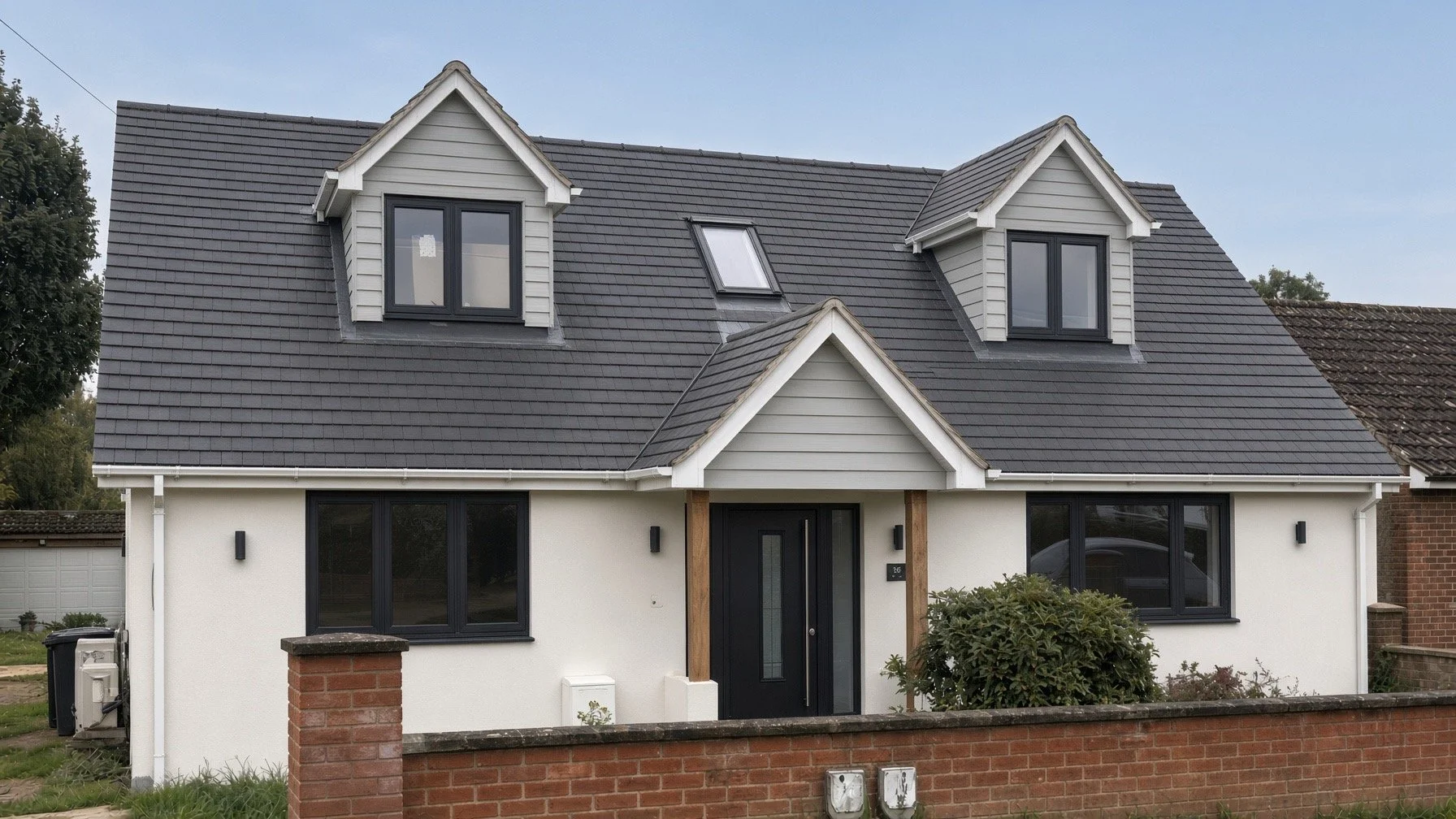 Front view of a modern house with a dark gray roof, two dormer windows, and a black front door, surrounded by a brick fence.