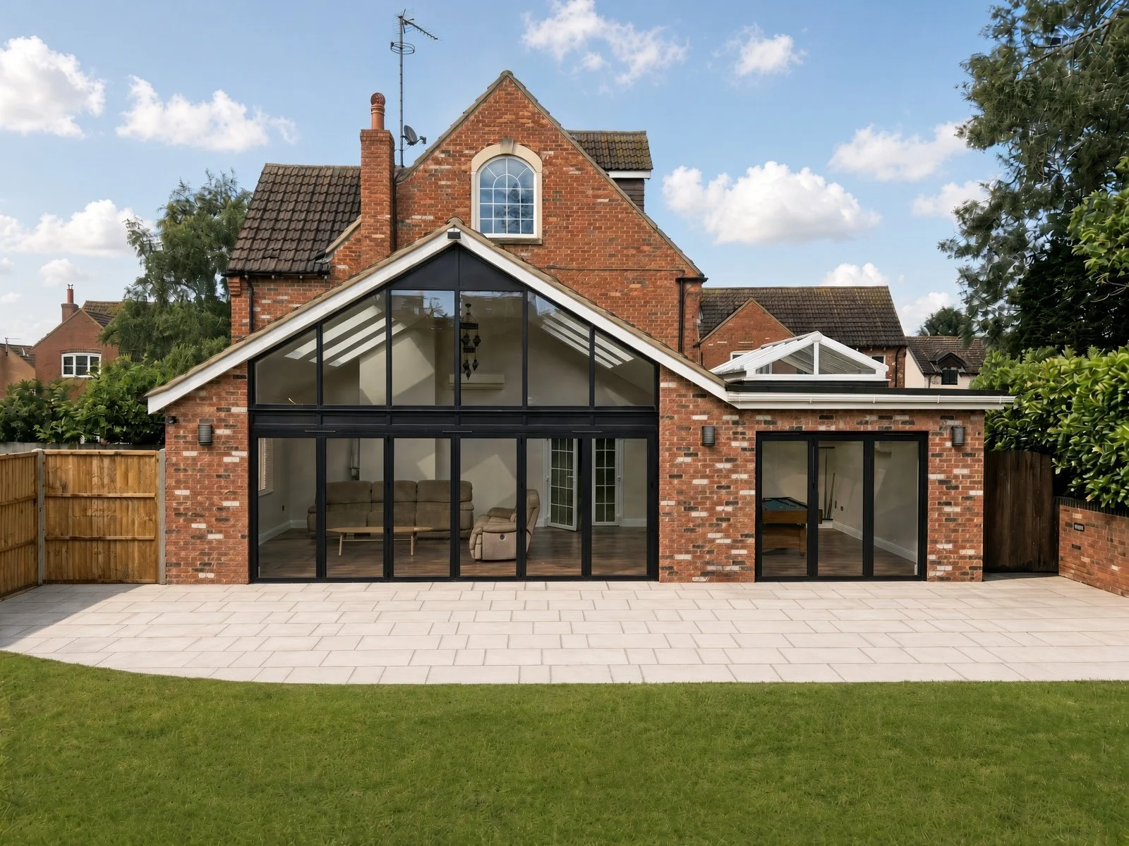 Backyard view of a brick house with a modern glass addition, featuring large sliding glass doors and windows, overlooking a paved patio and green lawn, with neighboring houses and trees in the background.