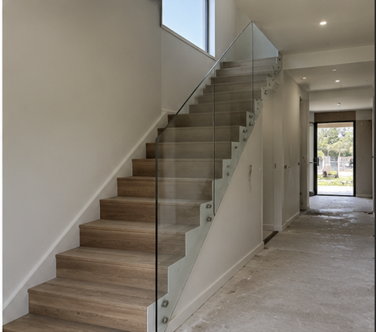 Interior view of a modern staircase with wood steps, glass railing, and a hallway leading to a door with a window showing trees outside.