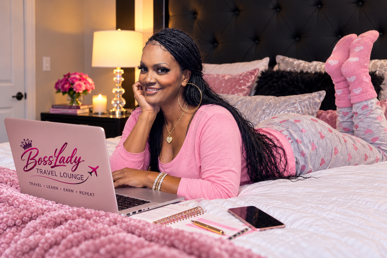 Woman lying on bed with pink themed bedding, working on a laptop. The laptop has a logo 'BossLady Travel Lounge' with a crown, airplane, and the words 'Travel, Learn, Earn, Repeat'. The woman is smiling, wearing pink pajamas with white hearts, large hoop earrings, and jewelry. There are pink flowers, a lamp, candles, and books on a side table in the background.