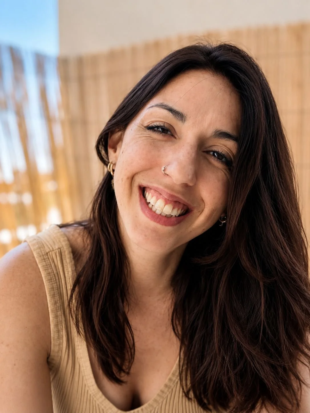 Portrait rapproché d’une jeune femme souriante aux longs cheveux bruns, portant un débardeur beige et un anneau au nez.