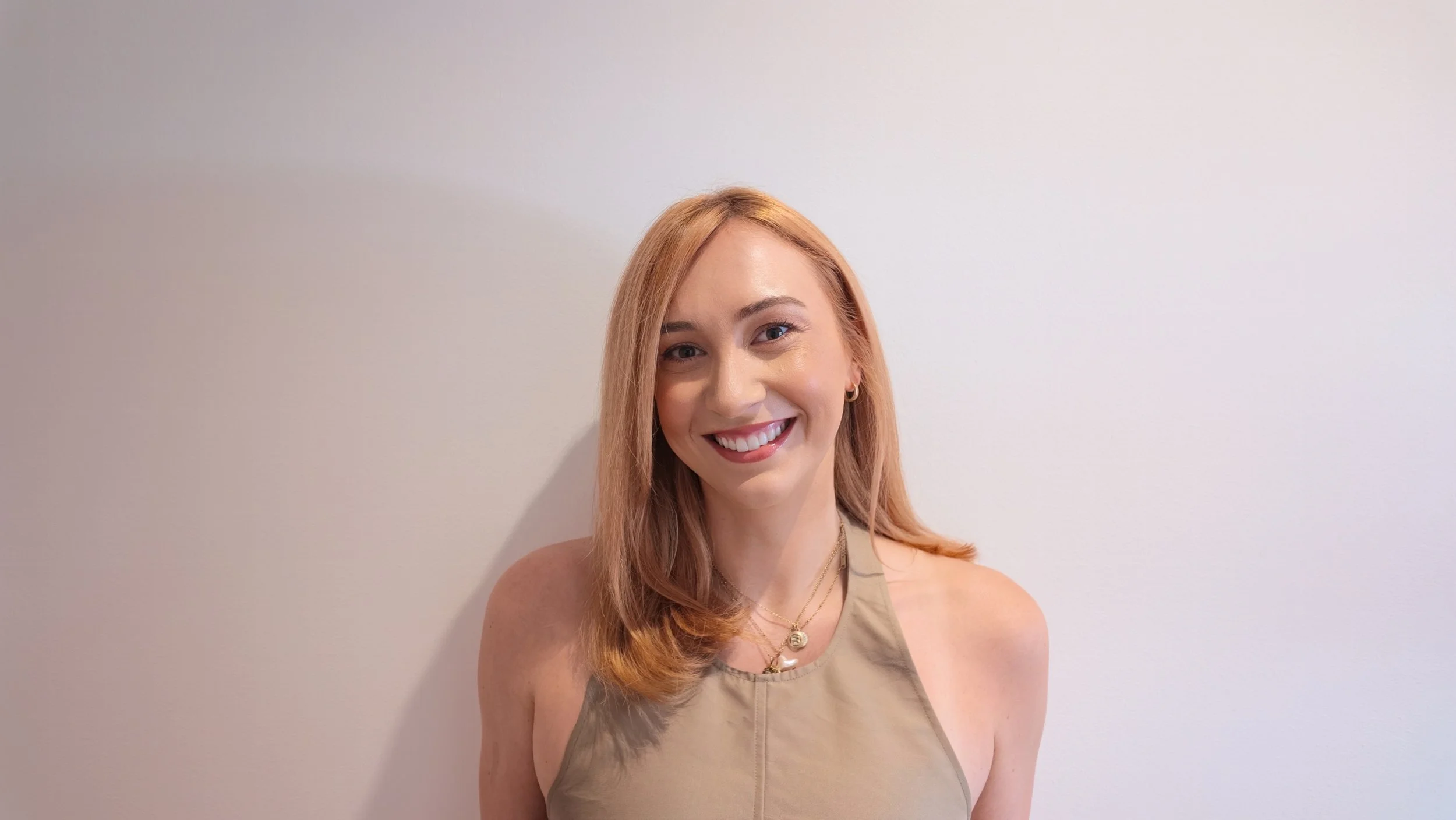 Smiling blonde woman wearing a beige sleeveless top and layered necklaces against a light wall