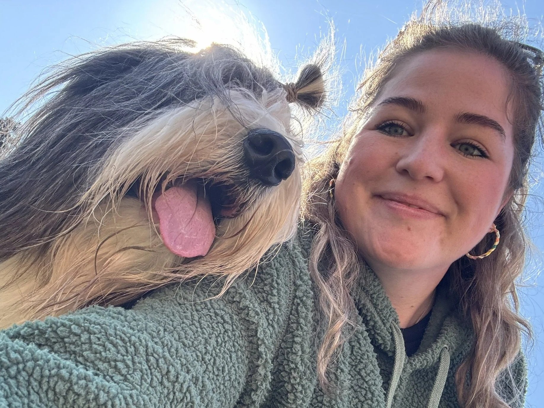 A woman with long hair and blue eyes taking a selfie with her dog outside on a sunny day. The dog has black and white fur, is panting with its tongue out, and is close to the camera, partially covering the woman's face.