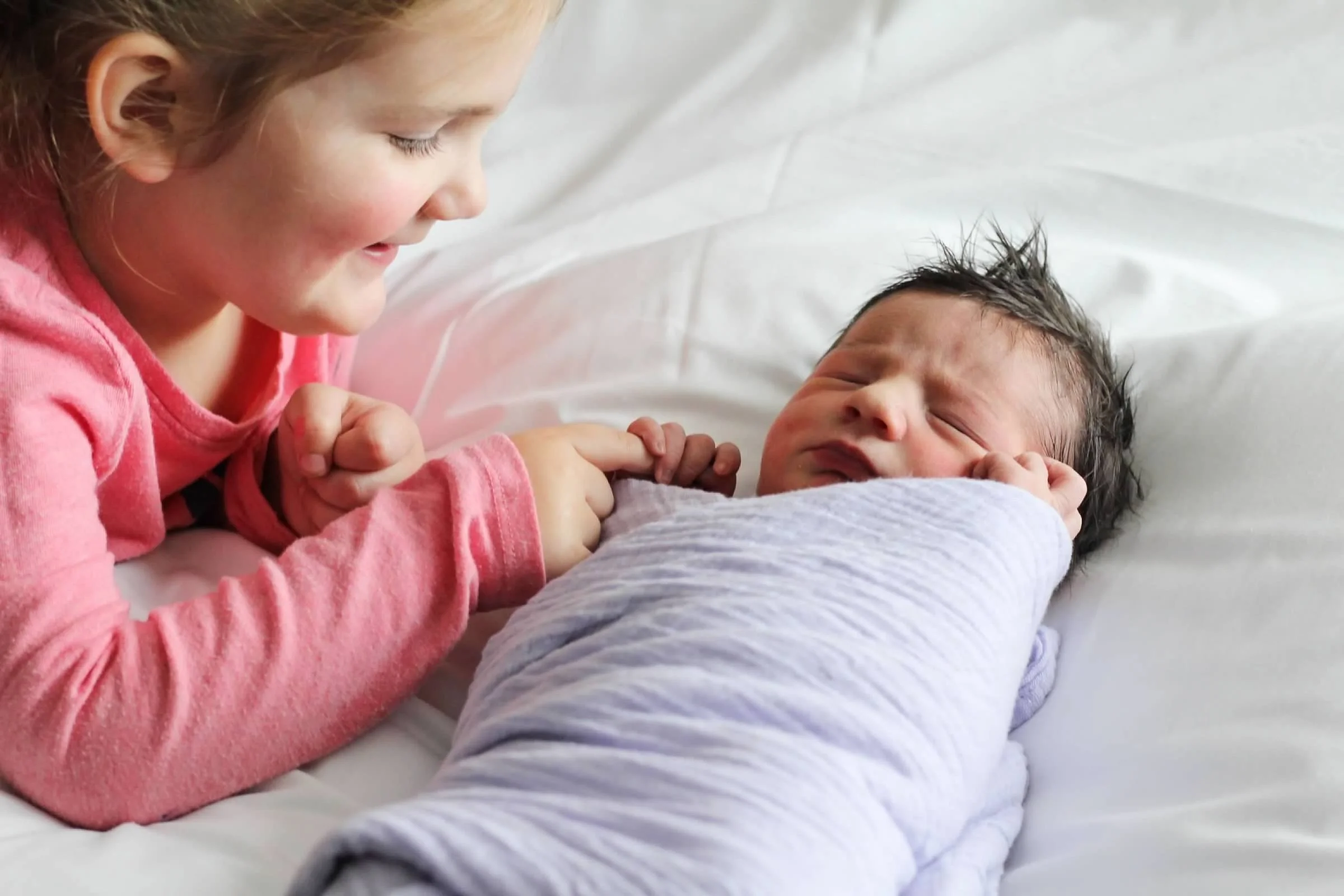 Young girl in pink pajamas gently holding a newborn baby wrapped in a lavender swaddle on a white bed