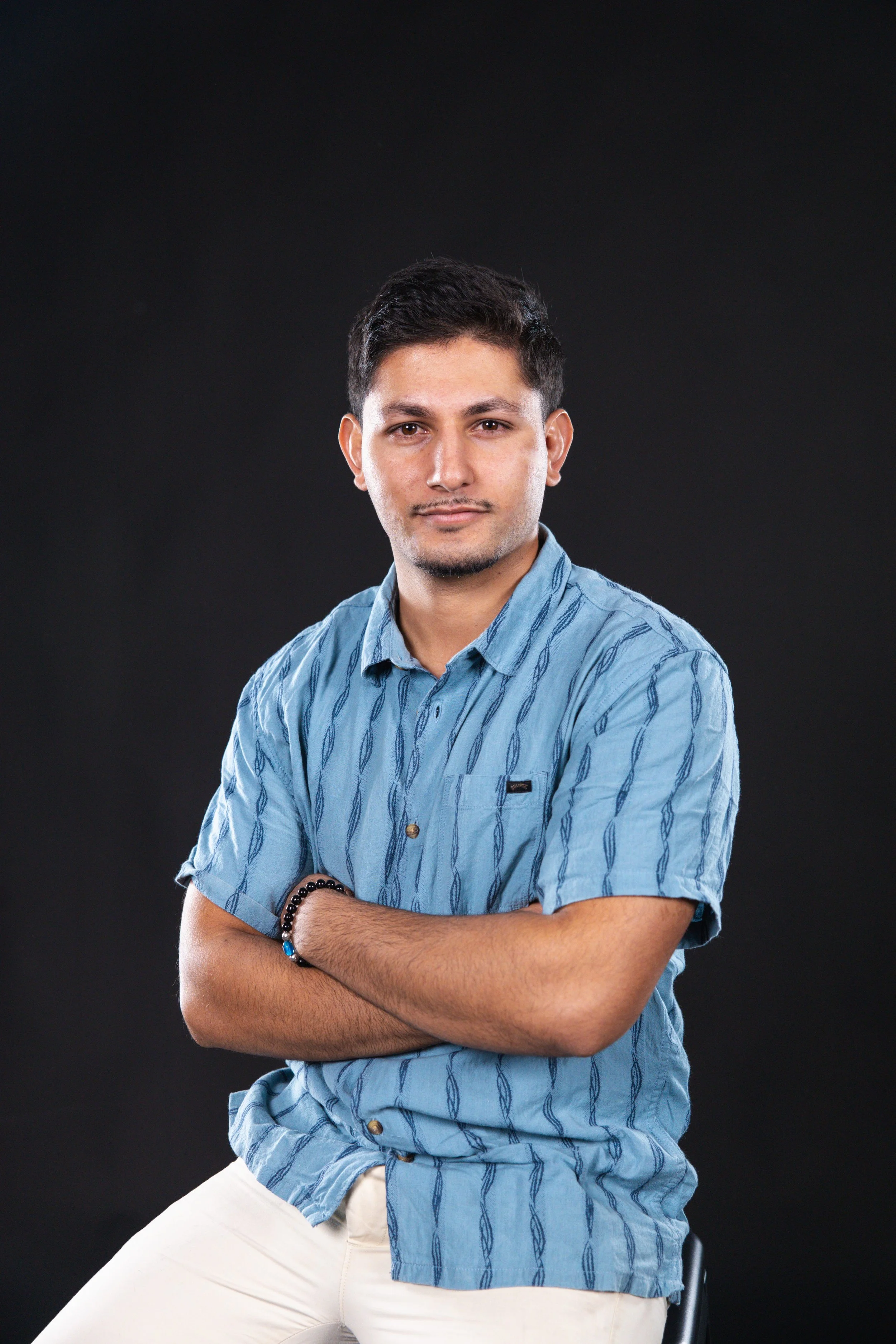 A young man with short dark hair, wearing a light blue patterned short-sleeve shirt and beige pants, sitting against a black background with arms crossed, looking at the camera.