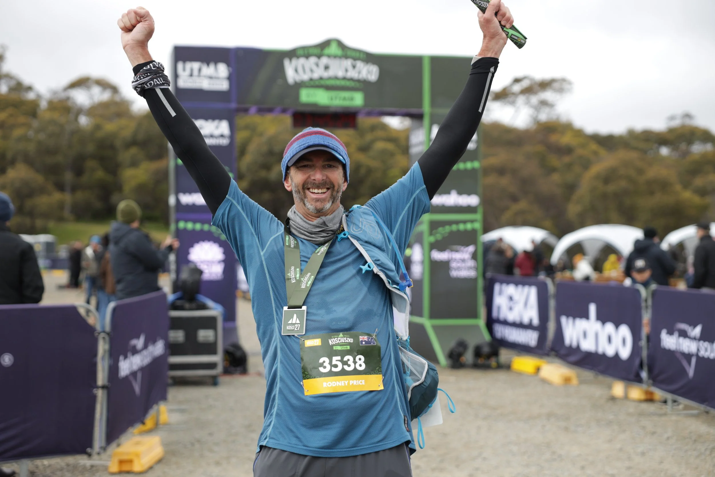 A man wearing a blue running shirt, black sleeves, and a cap stands at a finish line, smiling and raising both arms in victory. He has a race bib with the number 3538 and the name Rodney Price. The background shows a race event with several spectators and event banners.
