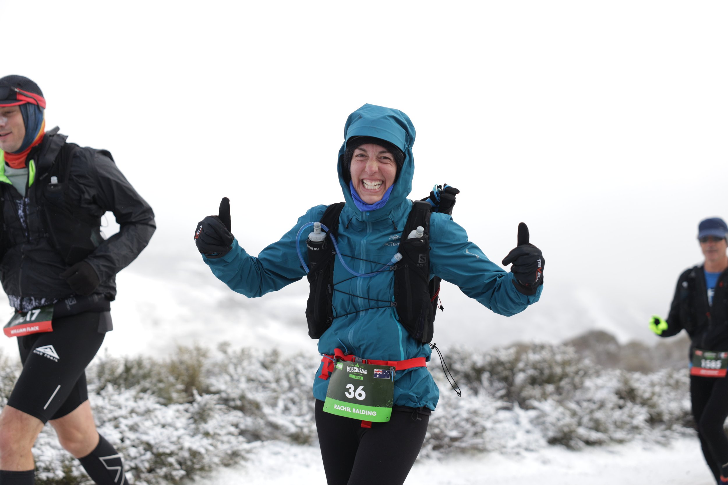 A woman in a blue jacket and black pants running in a snowy landscape during a race, smiling and giving thumbs up with both hands, wearing a race bib numbered 36.