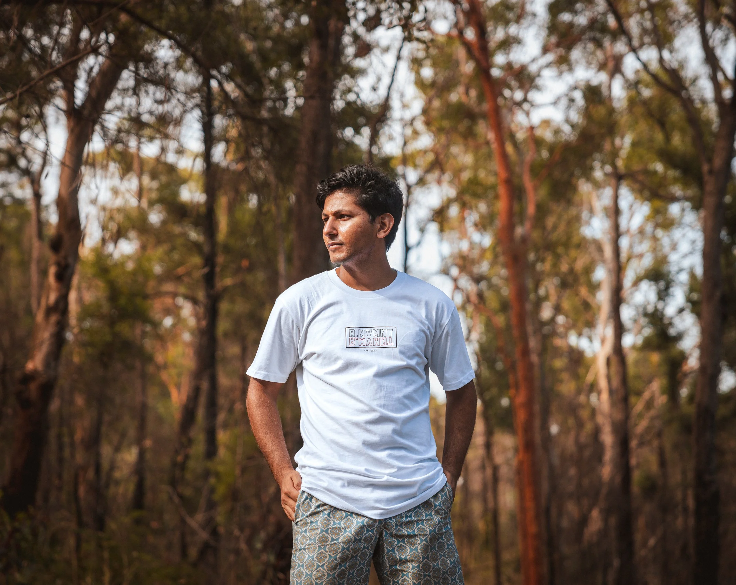 A man wearing a white t-shirt and patterned shorts standing in a forest with tall trees in the background.