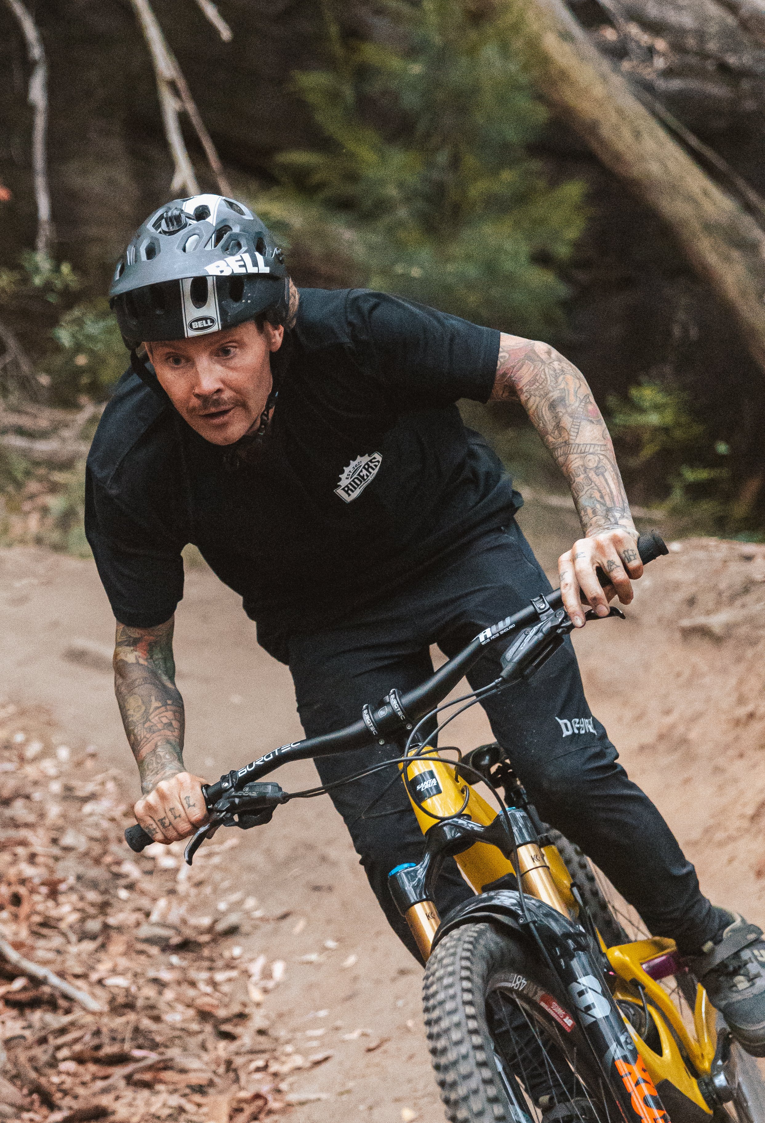 A man wearing a black helmet and black riding gear, with tattoos on his arms, is mountain biking on a dirt trail in a forest.