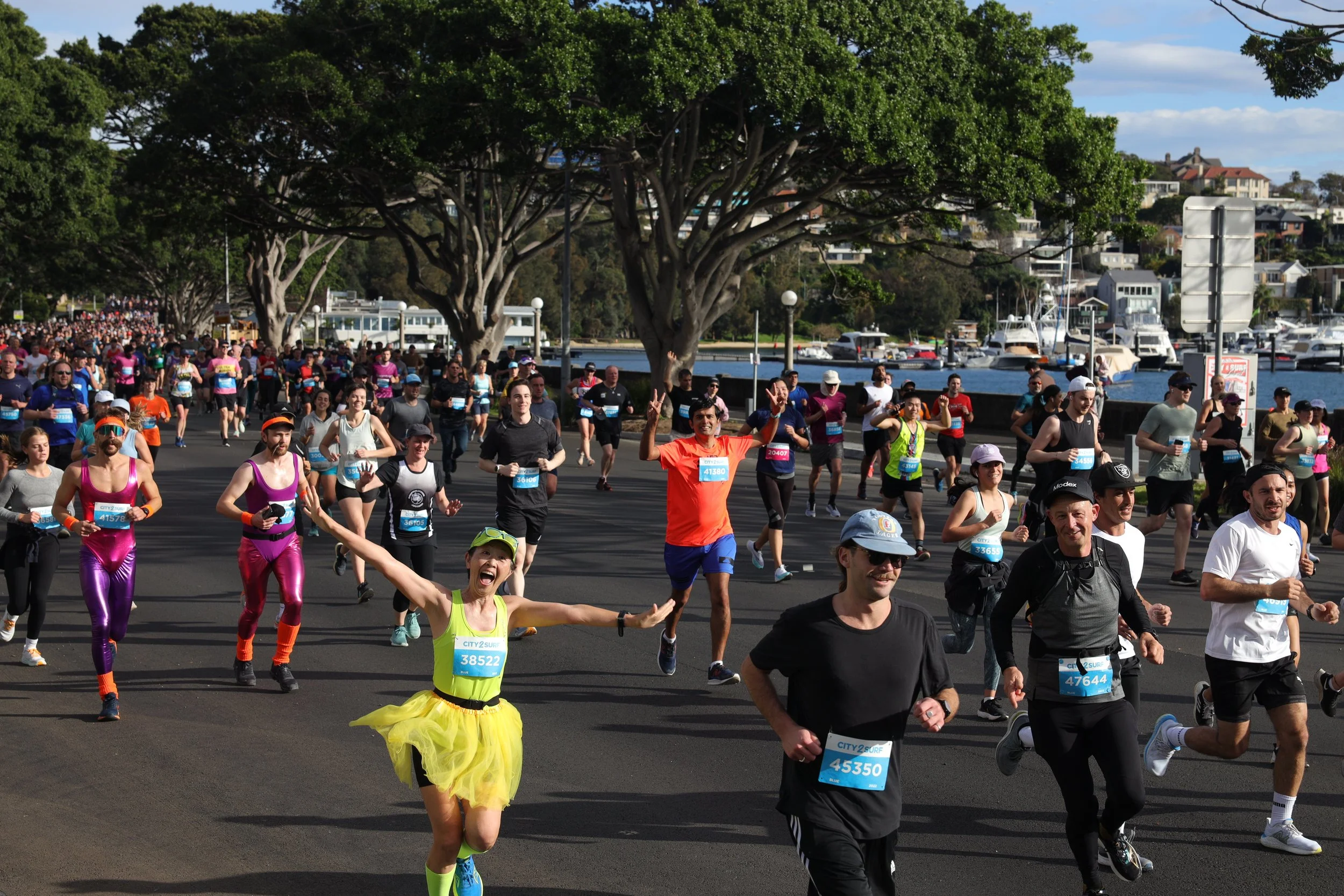 A large group of people running in a marathon along a waterfront, with some dressed in colorful costumes and athletic attire.