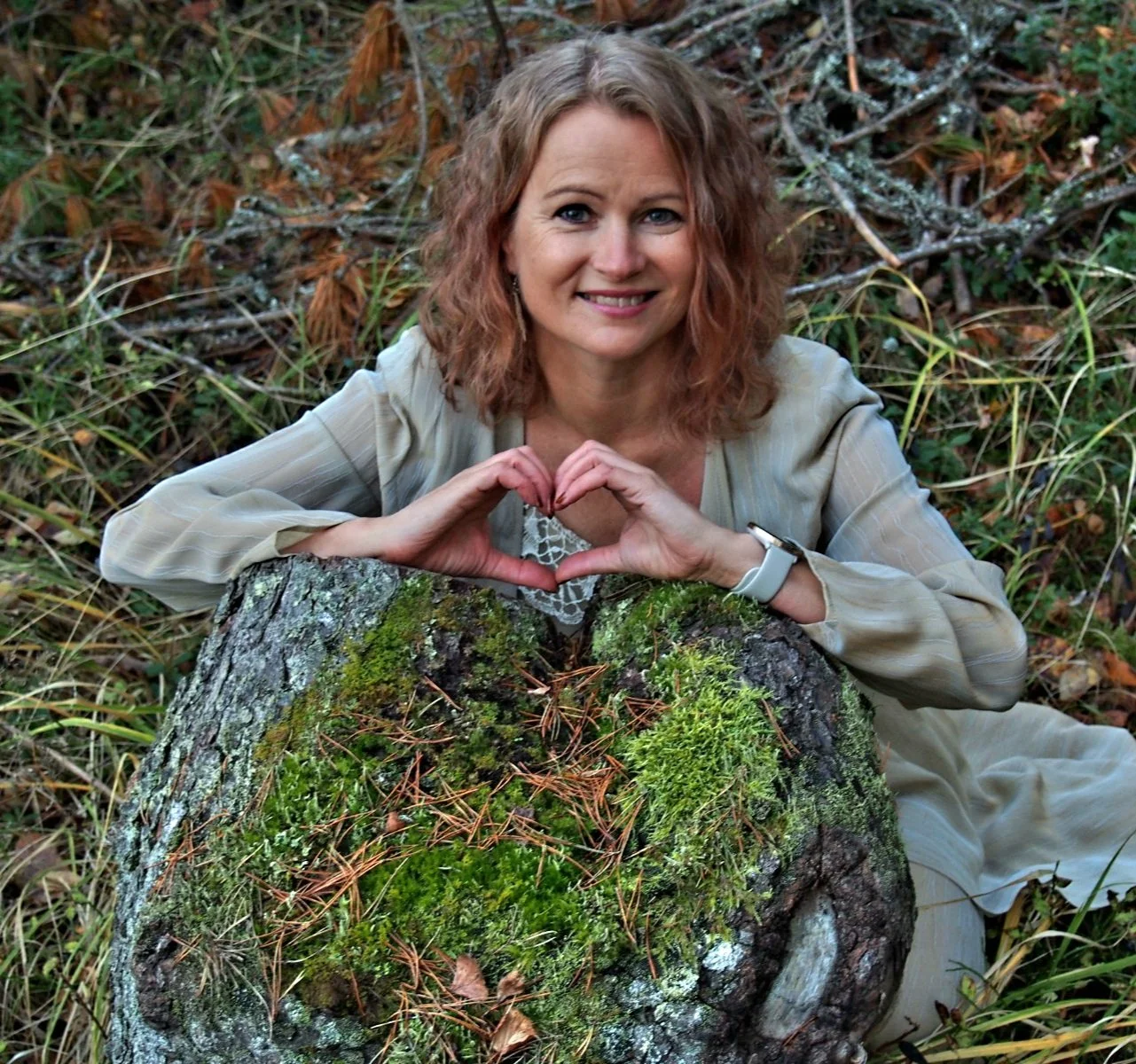 Hymyilevä nainen metsässä sammaleisen kannon takana, muodostaen käsillään sydämen.
