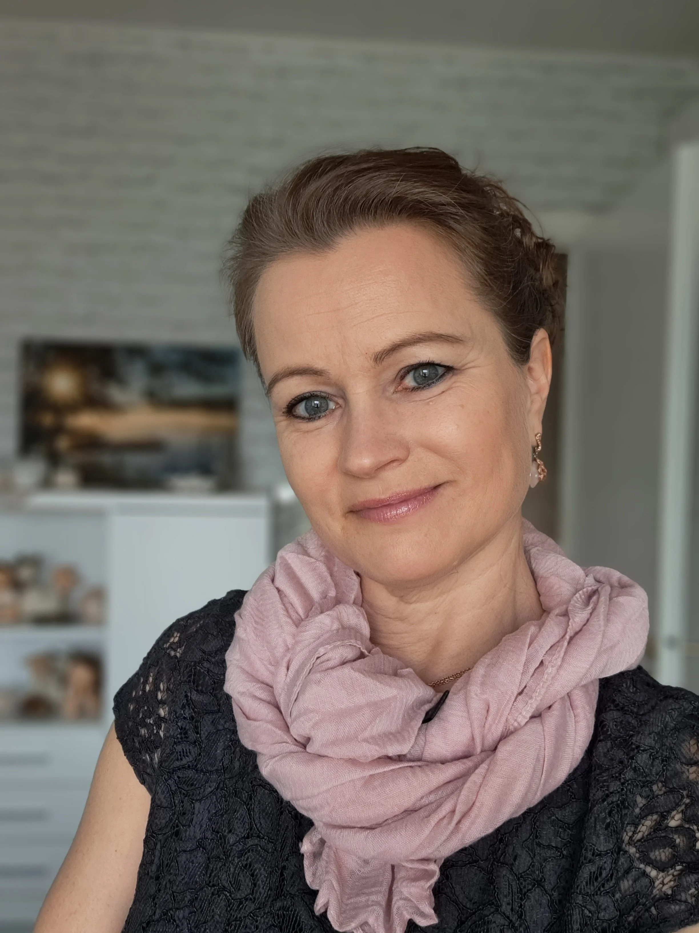 Lähikuva hymyilevästä naisesta, jolla on ruskeat hiukset, vaaleanpunainen huivi ja musta pitsipusero.