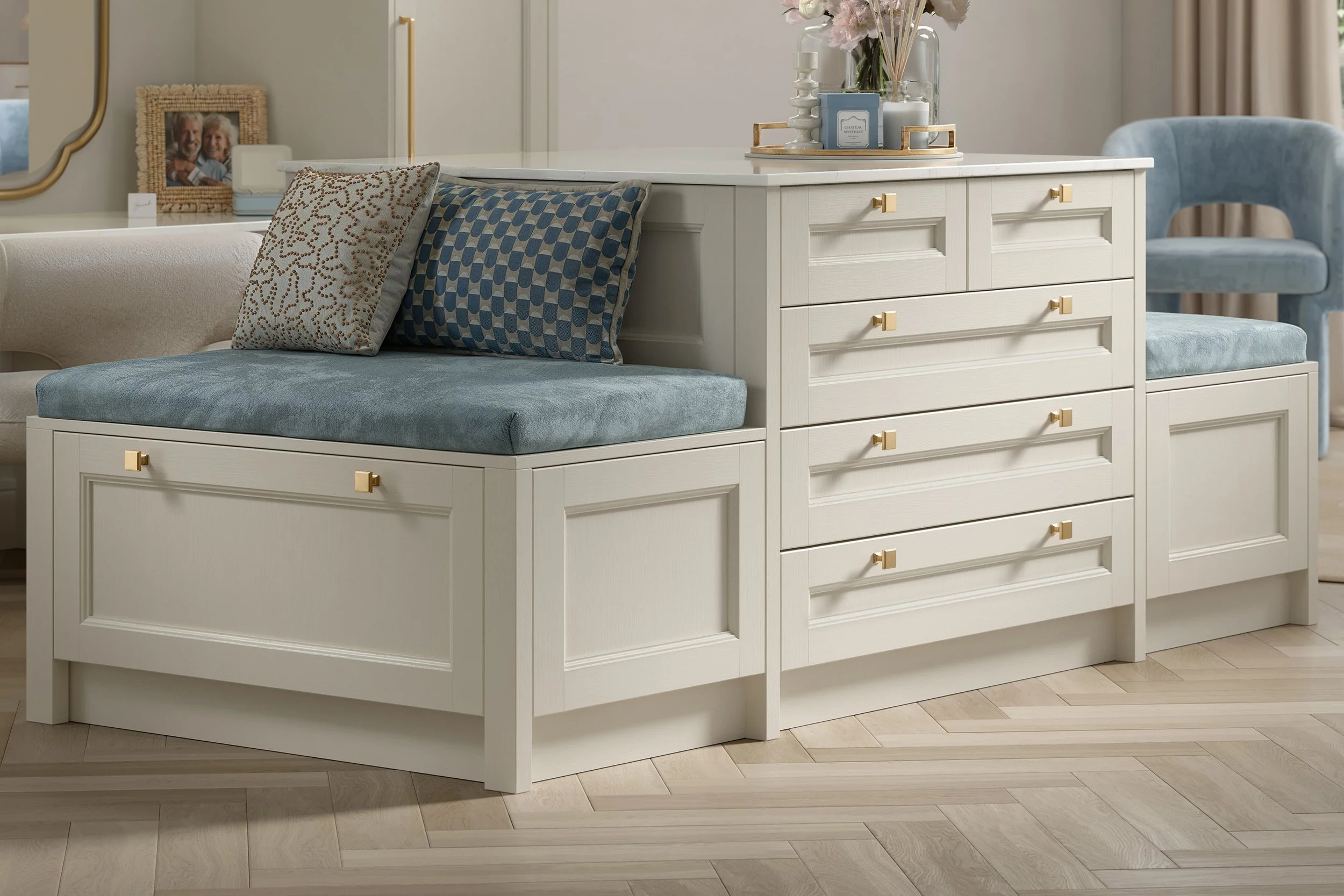 White built-in storage bench with blue cushion and patterned pillows beside a matching dresser, floral vase, and blue accent chair