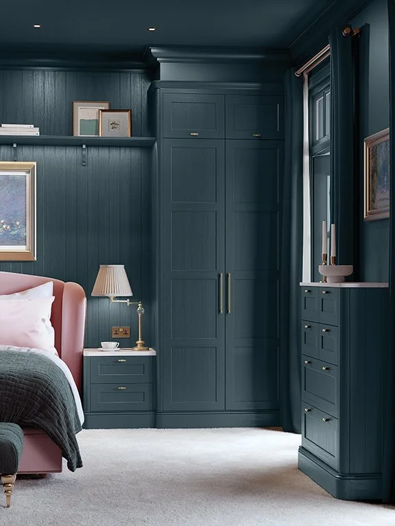 Elegant dark blue bedroom with paneled walls, built-in wardrobe, pink upholstered bed, nightstand, dresser, framed artwork, and soft carpeting