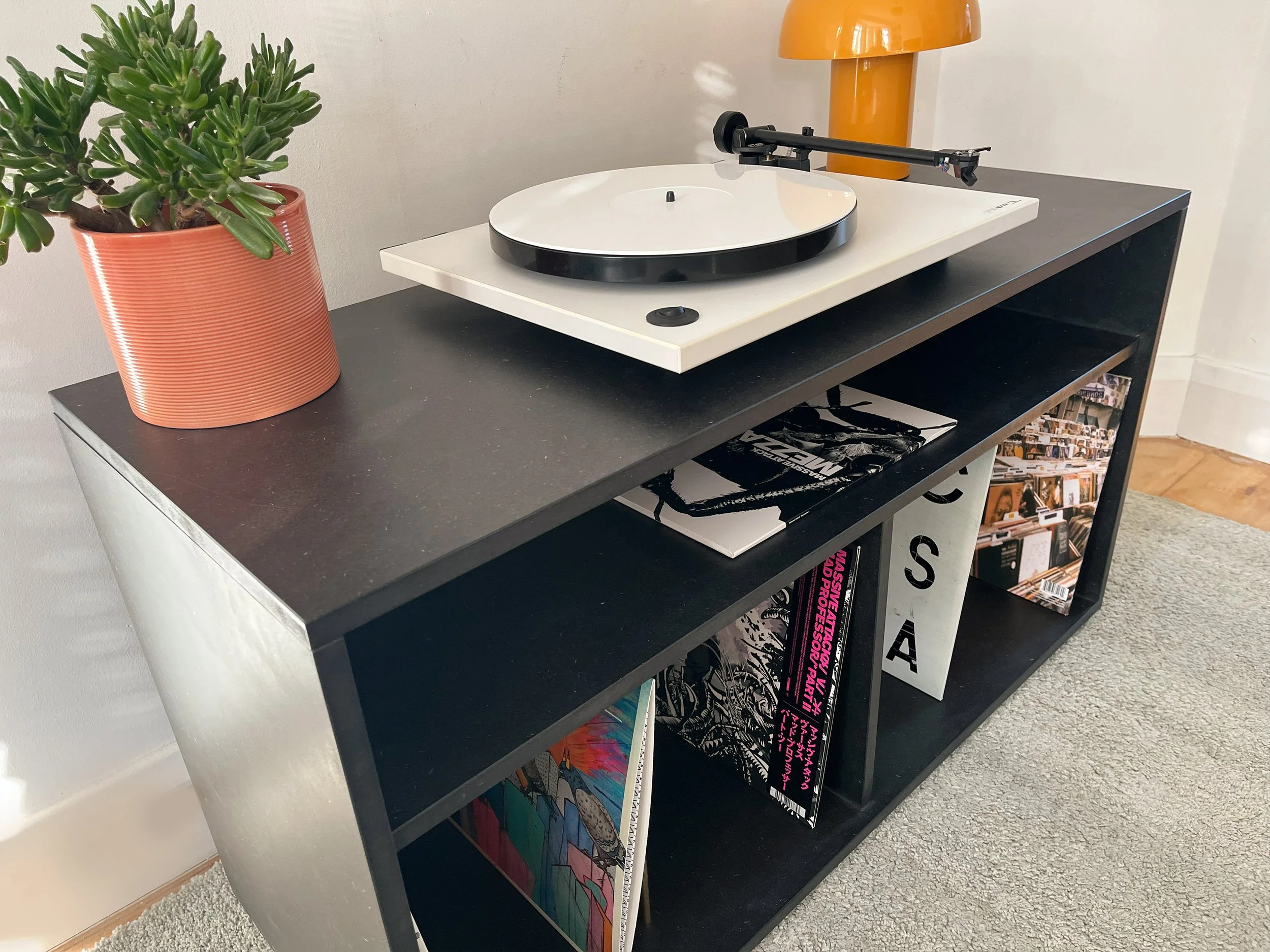 Black bookshelf with comic books and magazines, a white record player with a turntable on top, a potted succulent plant in an orange pot, an orange lamp, and a beige carpet flooring.