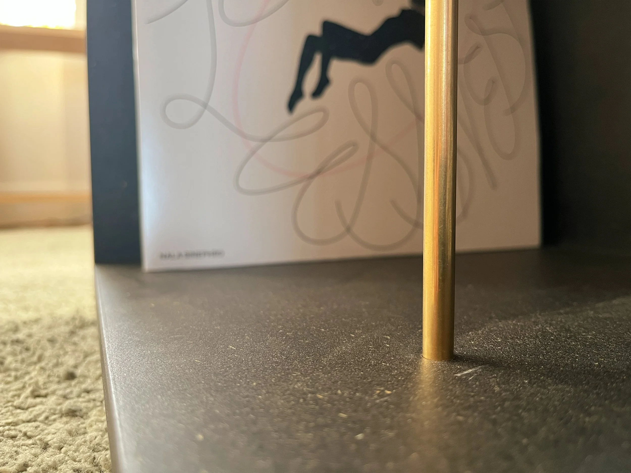 Close-up of a black hifi cabinet with a brass rod standing upright on the right side, with part of a box featuring abstract sketches and text in the background.