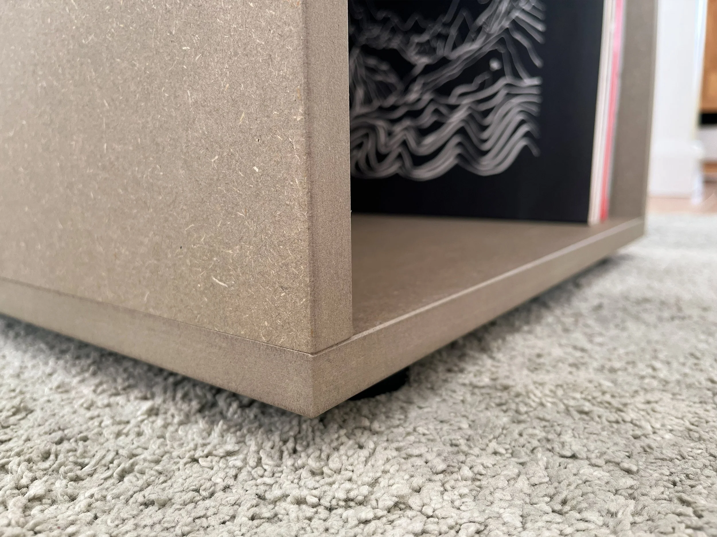 Close-up of a beige-colored piece of furniture or box with a black and white patterned object partially visible behind it, set on a gray carpeted floor.