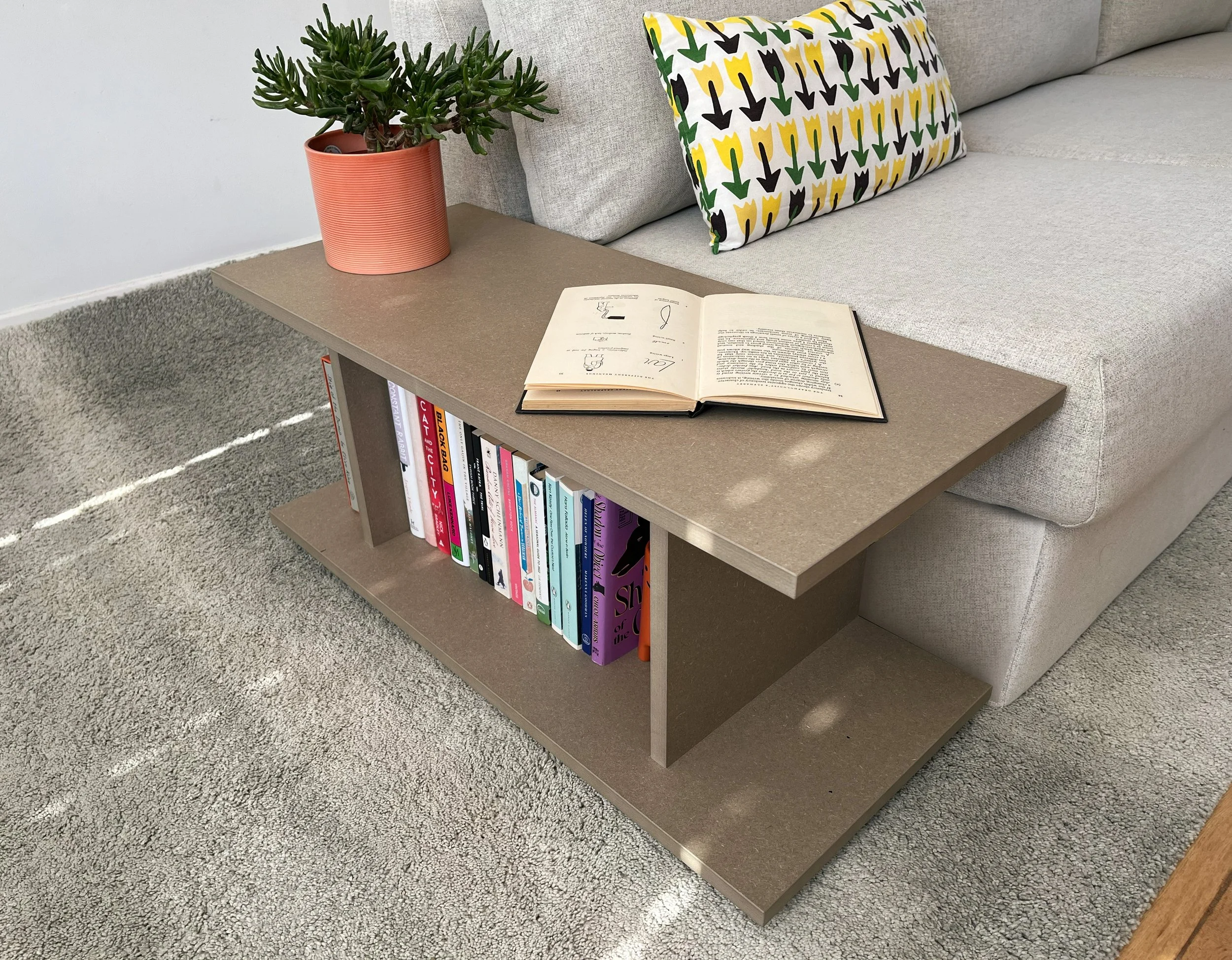 A coffee table with a potted succulent, an open book, and a bookshelf below, situated in a living room with a light-colored sofa and a decorative pillow with a black and yellow tulip pattern. The floor is covered with a textured light gray carpet.